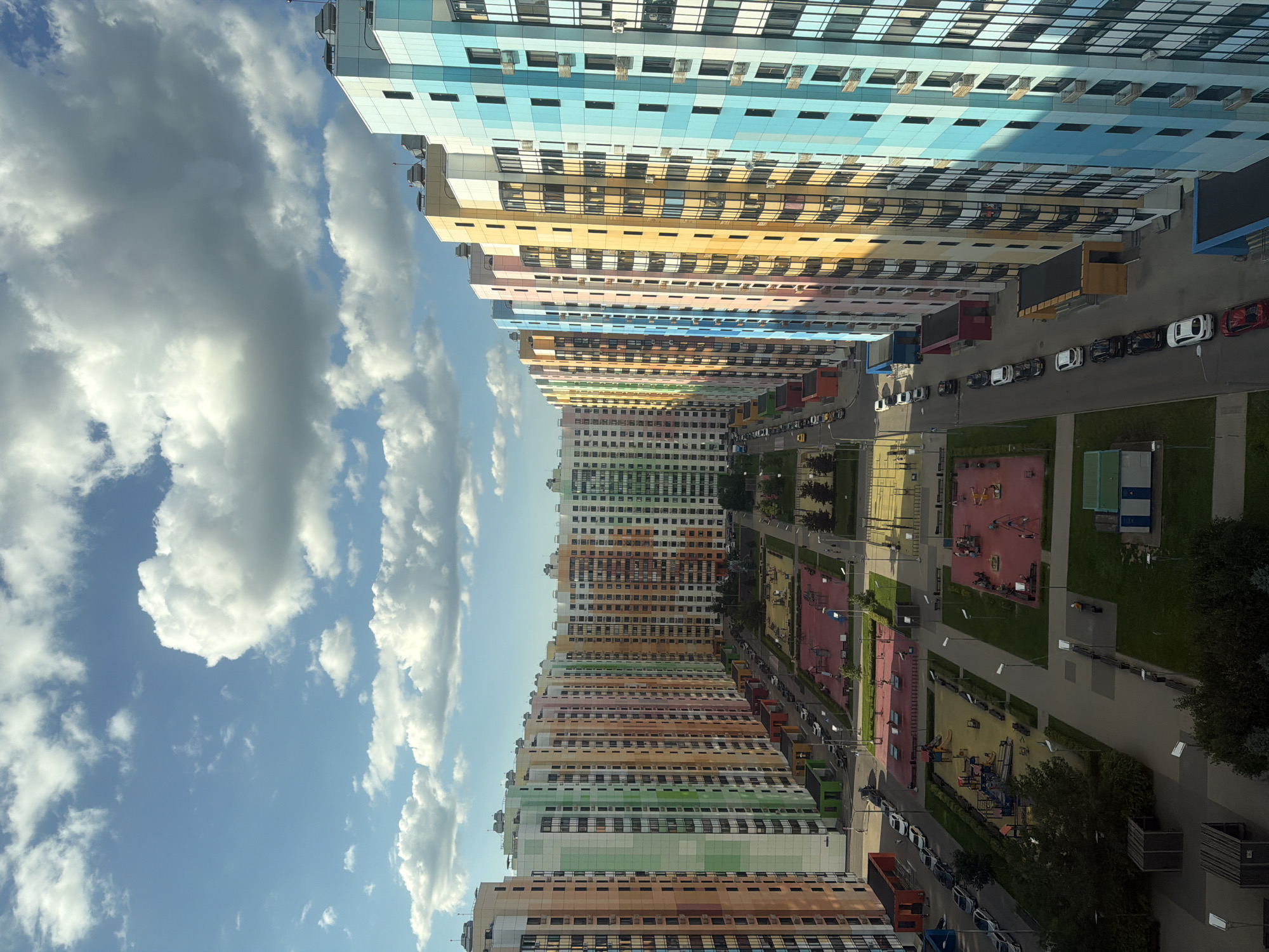 This is a high-angle, eye-level, medium shot of a modern urban residential complex with numerous high-rise buildings. The buildings are colorful, with facades painted in shades of blue, yellow, pink, orange, green, and peach. They are arranged in a U-shape, creating a central courtyard area. The courtyard contains several children's playgrounds with red and yellow surfaces, swings, and climbing structures, as well as benches and green spaces with trees. A paved pathway runs through the courtyard, connecting the buildings and playgrounds. Cars are parked along the street adjacent to the buildings and on a smaller street within the complex. The sky is blue with scattered white, puffy clouds. The scene is brightly lit, suggesting it is daytime. The overall impression is one of a well-maintained, modern, and vibrant residential neighborhood.