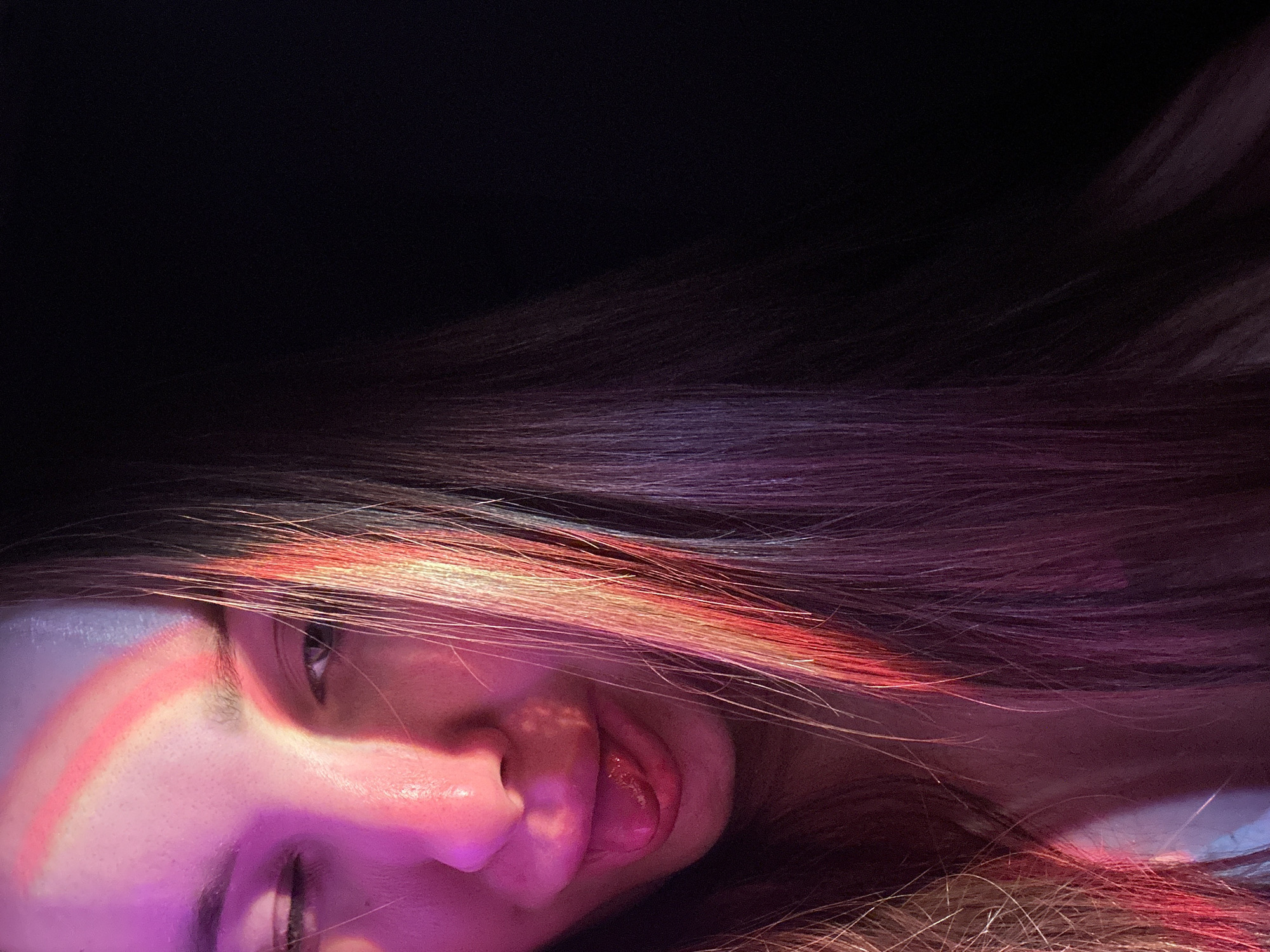 The image shows a person lying down, with their face and hair illuminated by a pink and red light. The background is dark. The person's tongue is slightly out. There is no information to suggest the location is Chaikovsky, Russia.