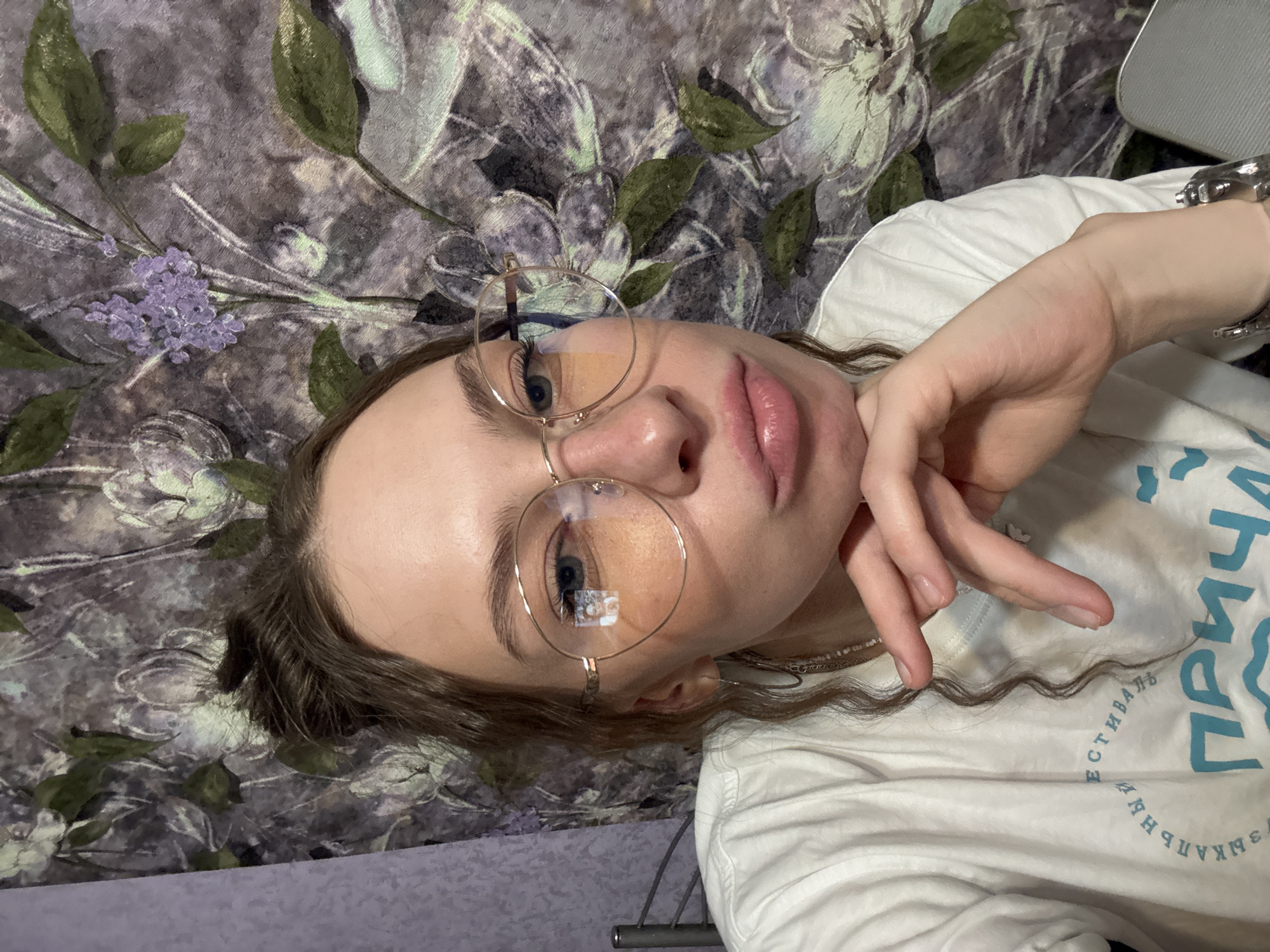 A young woman with her hair in a messy bun and wearing large round glasses, a white t-shirt, and hoop earrings, rests her chin on her hand. Her mouth is pursed, and she is looking at the camera. The background is a floral wallpaper with purple and green tones. The image appears to be a selfie taken indoors. There is no visible indication of the location being in the city of Perm, Russia.