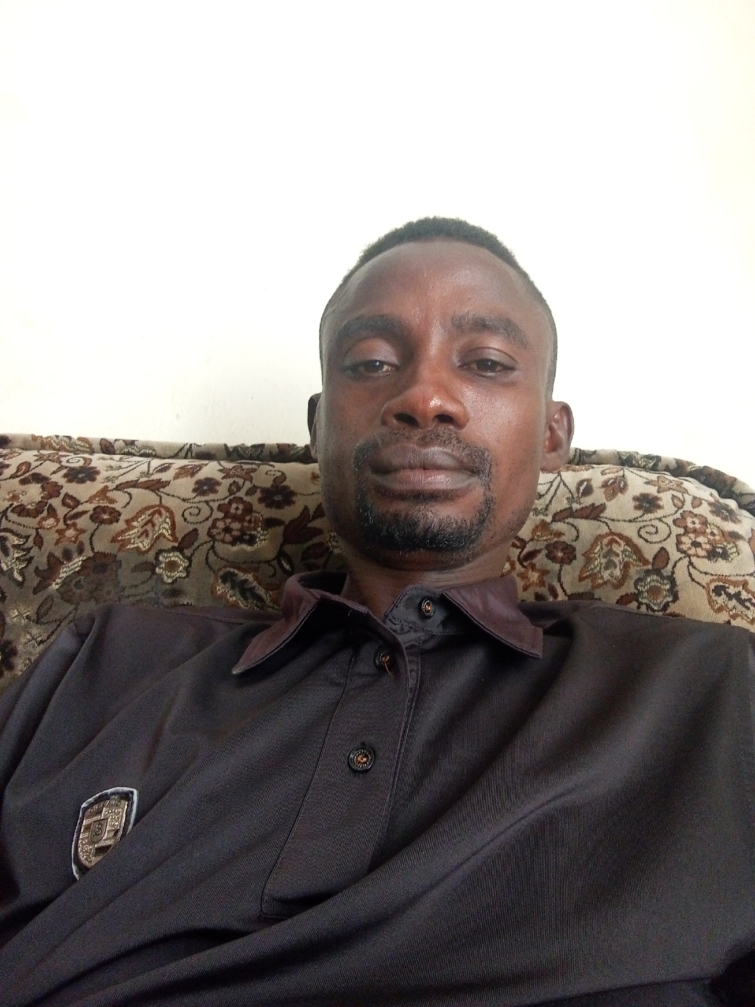 A man is sitting on a patterned couch, wearing a dark button-down shirt with a small emblem on the left chest. The location is Unknown, Unknown. The background is a plain white wall.