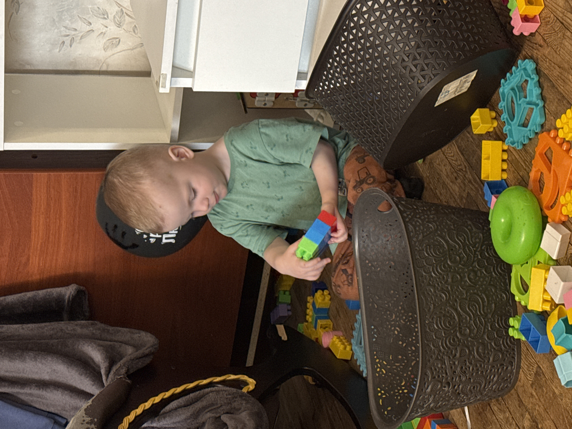 A child plays with toys on the floor next to two black plastic baskets. The child wears a green shirt and brown pants and is building with blue, green, and red blocks. The surrounding floor is covered in colorful plastic blocks of various shapes. The location cannot be determined based on the visual information.