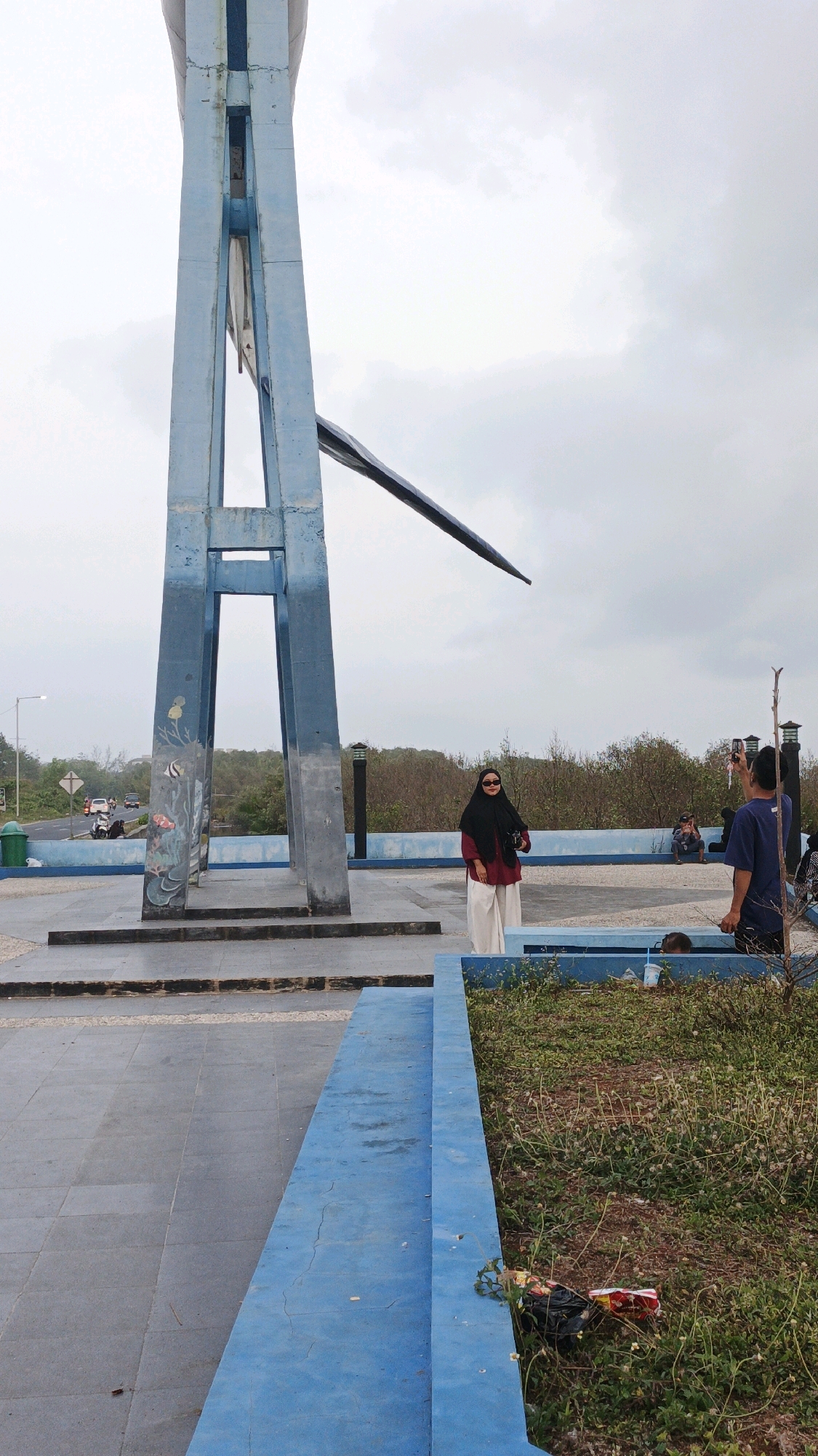 A woman wearing a black hijab, maroon top, and white pants stands near a tall, blue, abstract sculpture. She is facing the camera and smiling. To her right, a man in a blue shirt holds up a phone, appearing to take a photo of her. The sculpture is located in an outdoor public space, possibly a park or monument area, with paved walkways and planted areas. In the background, trees and a road with passing vehicles are visible. The sky is overcast. The scene appears to be in Pangandaran, Indonesia, suggested by the presence of coastal vegetation and the general atmosphere.