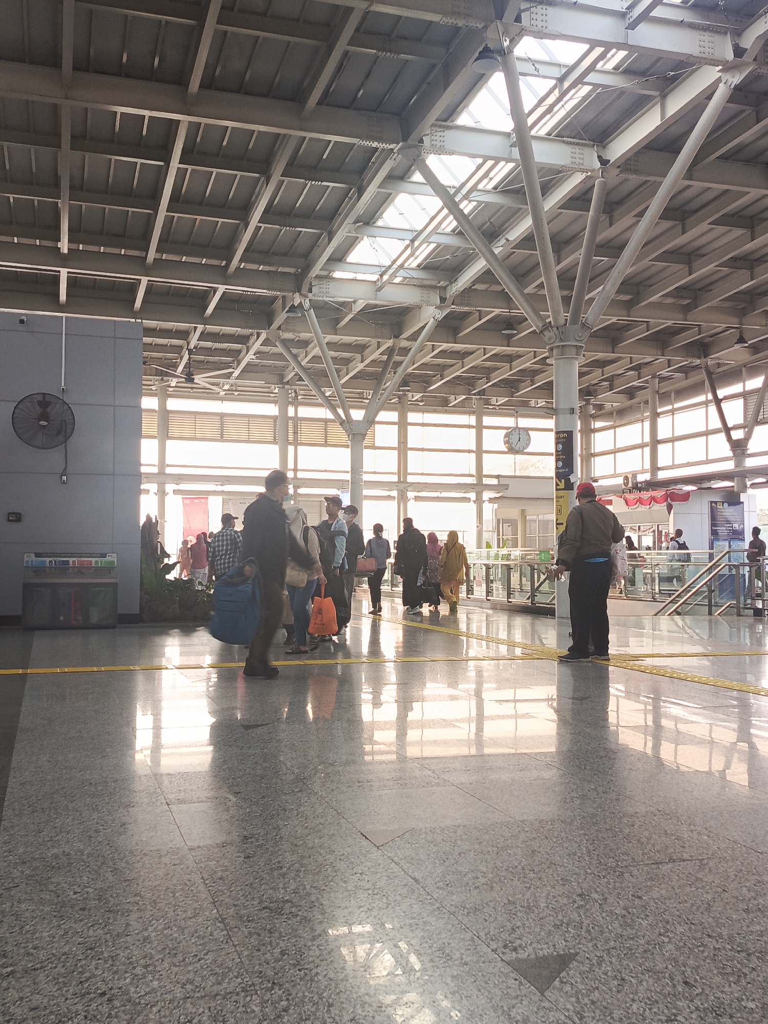 The image captures the interior of a public transport station, likely a train or bus station, in East Jakarta, Indonesia. The scene is characterized by a high, industrial-style ceiling with exposed metal beams and translucent panels that allow natural light to filter through, creating a bright and airy atmosphere. The floor is a polished, speckled grey material that reflects the light and the people moving across it.
Several individuals are visible throughout the station. In the foreground, a person wearing a dark jacket and a red cap stands with their back to the camera, facing towards a set of stairs. To the left, a group of people are gathered, some with luggage, appearing to be waiting. Among them, a person in the foreground is carrying a large blue bag. Further into the station, more individuals are seen walking or standing, some of whom are women in traditional Indonesian attire, including headscarves.
Key objects in the scene include a large, round clock mounted on a pillar, a black industrial fan mounted on a grey wall to the left, and trash receptacles near the fan. Yellow tactile paving lines are visible on the floor, indicating pedestrian pathways. The background shows glass barriers, staircases, and advertisements or information displays. The overall impression is of a busy transit hub, with natural light playing a significant role in illuminating the space.