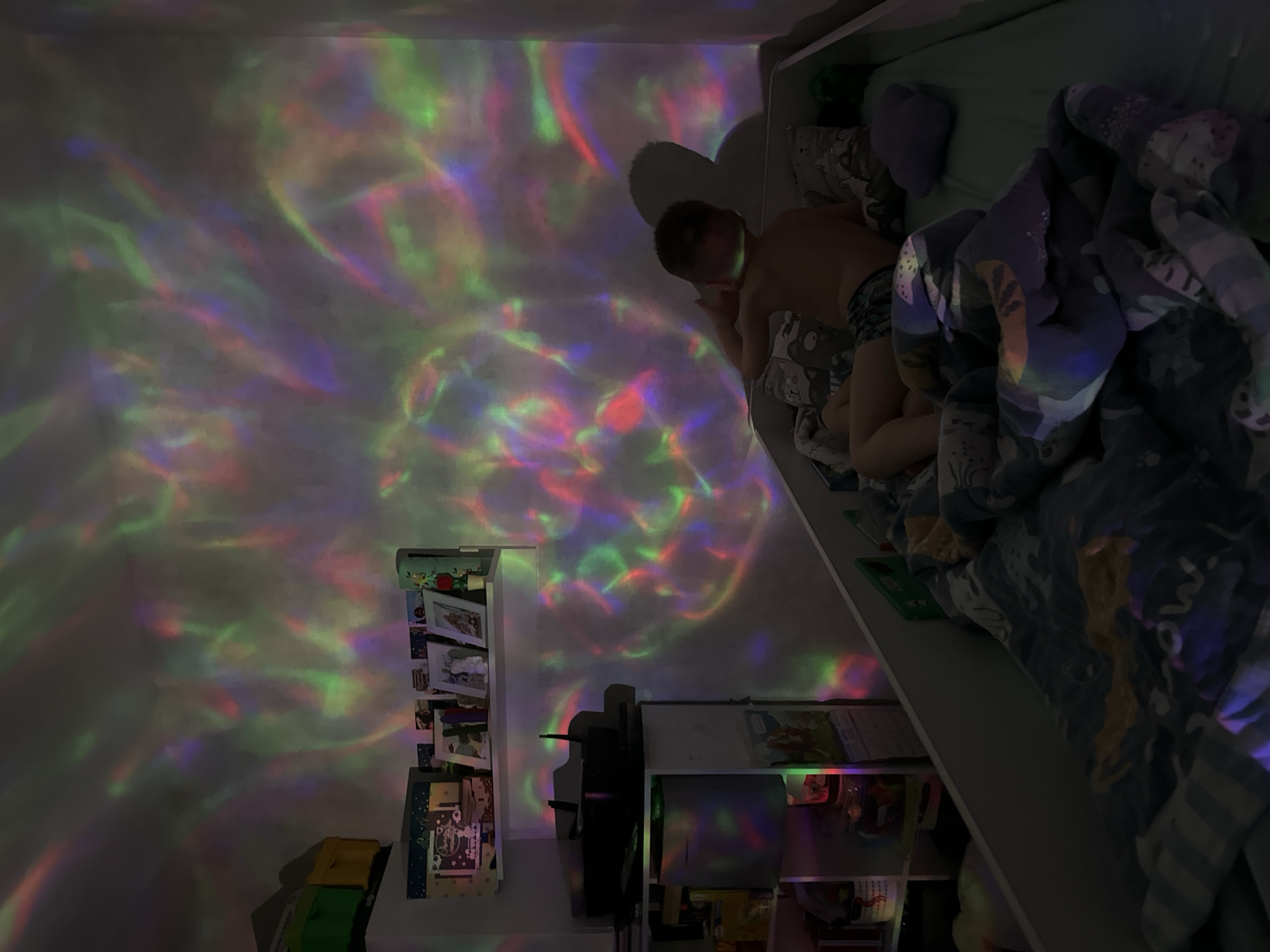 This media file captures a nighttime scene in a bedroom in Tomilino, Russia, illuminated by a colorful light projector. The image is oriented with the bed appearing vertically on the right side. A young child, partially visible, lies in a bed with a light blue/grey patterned duvet and a purple pillow. The child is wearing dark patterned shorts and appears to be resting or sleeping.
The most striking feature of the scene is the dynamic display of swirling, nebulous patterns of red, green, blue, and purple light projected onto the walls and ceiling of the room, creating an ethereal and calm atmosphere. The room itself is dimly lit, emphasizing the projected lights.
Below the bed, and along the left side of the frame (which appears as a wall), are white shelving units filled with various items. These include framed photographs, books, and what appear to be toys or decorative objects. Another shelf unit is visible in the lower right foreground. The bed seems to be a bunk bed or a high sleeper, elevated above some of the shelving.
The activity depicted is that of a child resting in a uniquely lit bedroom, possibly as part of a bedtime routine or simply enjoying the projected lights. No legible text is discernible on any of the objects within the image. The overall mood is peaceful and imaginative.