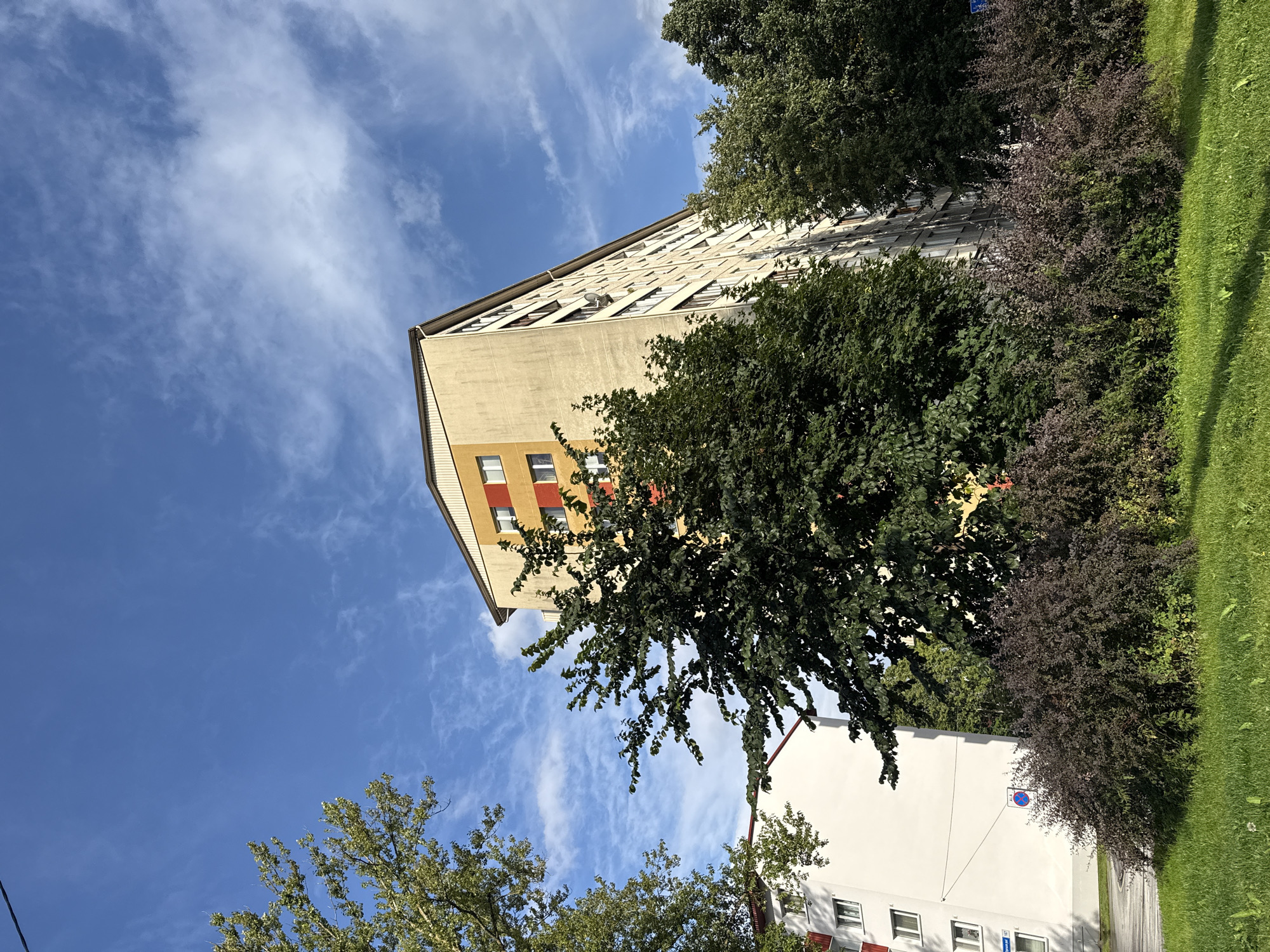 The image shows a view of apartment buildings in Tallinn, Estonia, on a sunny day with a blue sky and scattered clouds. In the foreground, there is a patch of green grass and a cluster of dark-leafed bushes. Behind the bushes, a large, light-colored apartment building dominates the center of the frame. This building has a beige facade and a sloping roof painted yellow. Sections of the building are accented with red and yellow panels around the windows. To the left of this building, a smaller, white apartment building with a red-tiled roof is partially visible. Lush green trees frame the apartment buildings, with their leaves creating shadows on the facades and the ground. The overall scene depicts a residential area in an urban setting.