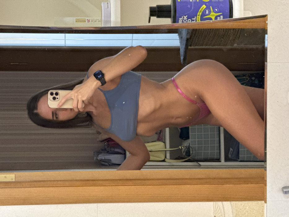 A mirrored image shows a slender woman in a blue sports bra and pink thong. She holds a phone, taking a selfie. Her toned physique is visible. In the background, bathroom items and storage can be seen. The mirror's wooden frame is partially visible.