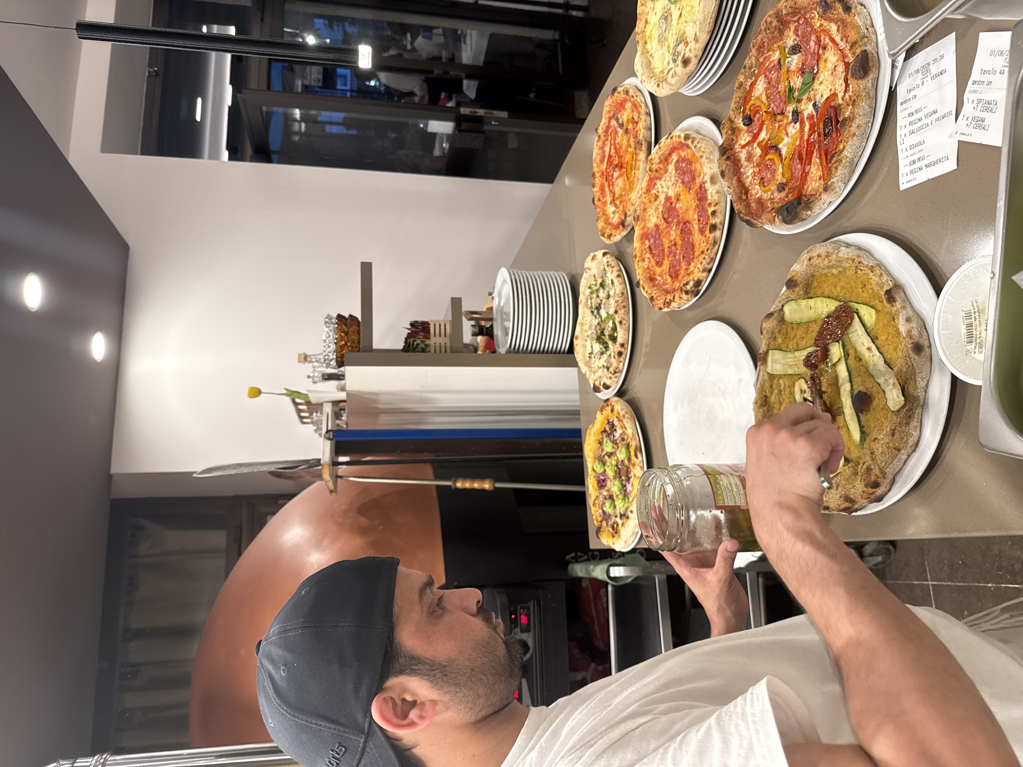 A man wearing a black baseball cap and a white t-shirt is standing in what appears to be a pizza restaurant, possibly in Trento, Italy, as suggested by the text on the receipt visible in the lower right. He is in the process of preparing or garnishing a pizza with a fork, holding a glass jar of what looks like olive oil in his left hand. In front of him and to his right, there are several pizzas already prepared and arranged on a counter. These pizzas exhibit various toppings, including what appear to be salami, vegetables like zucchini and peppers, and cheese. Behind the man, a large, domed pizza oven is visible. The environment suggests a commercial kitchen setting with stacked plates and lighting fixtures overhead. The overall scene captures a moment of food preparation in a culinary establishment.