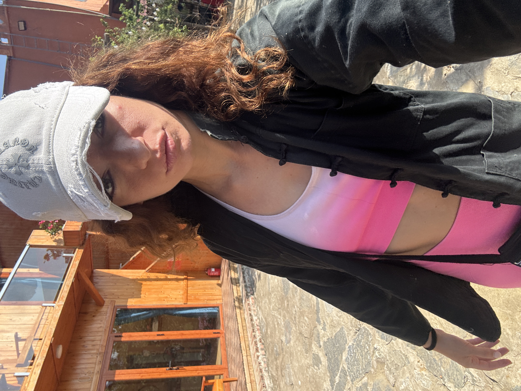 A woman wearing a gray cap, a black jacket, and a pink and white sports bra and leggings stands outdoors on a paved surface. Buildings are visible in the background. The location is possibly in Bắc Hà, Vietnam.