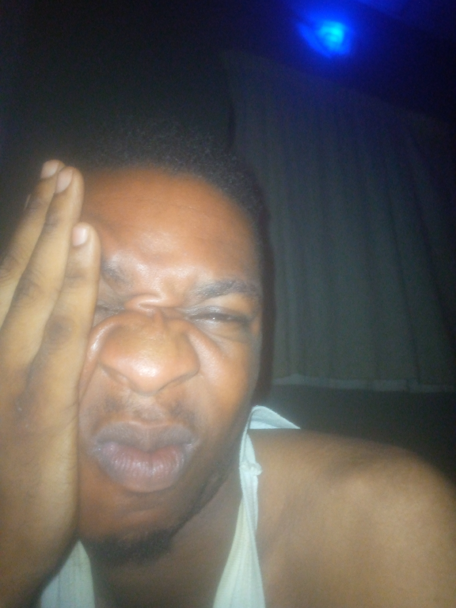 A close-up indoor shot features a dark-skinned person pressing the palm of their hand against the left side of their face. Their face is contorted, with eyes squinted shut, nose scrunched, and lips pursed. The person is wearing a white sleeveless garment. The background is dark and somewhat blurry, with what appears to be a section of white curtain visible on the right side. A blue light source is visible near the top of the frame. The location is described as Unknown, Unknown.