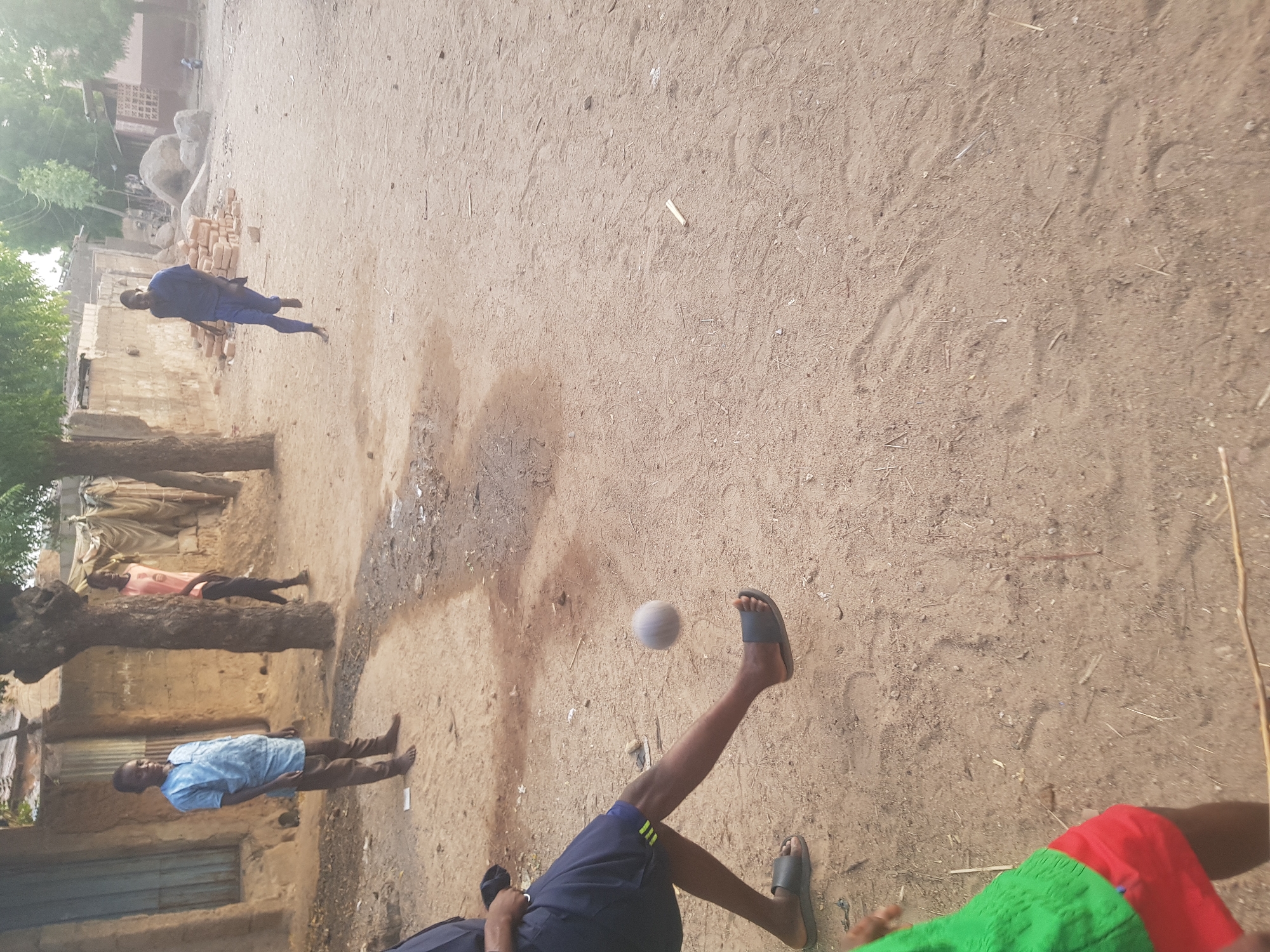 The image depicts an outdoor scene in Dutsinma A, Nigeria. The ground is sandy and uneven. Several people are present, with two barefoot men standing near a dilapidated structure made of mud bricks. Another man in blue clothing is lying on the ground further away. In the foreground, a person wearing shorts and sandals is kicking a white ball. The overall impression is a candid shot of everyday life.