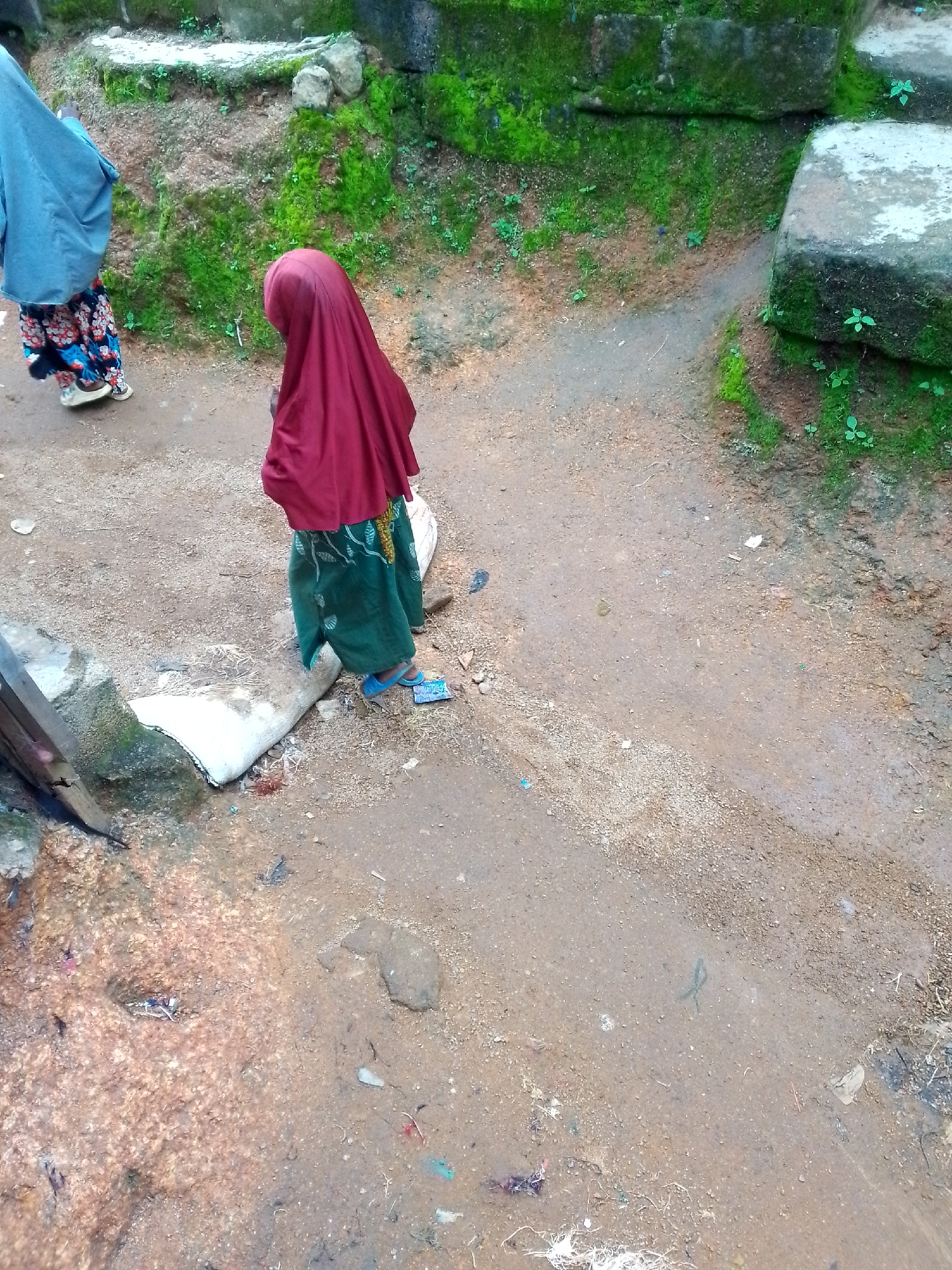 Two individuals are visible in the frame, outdoors, standing on a dirt path. The person on the left is wearing a light blue head covering and a patterned skirt, with their back to the viewer. To the right of this individual, a second person is seen from the back, wearing a dark red head covering and a green skirt adorned with a white leaf pattern. This person is wearing blue flip-flops. The ground is composed of dirt and small pebbles, with patches of green moss and vegetation visible on raised earthen mounds and concrete structures in the background. Some discarded items, including a white cylindrical object and various pieces of debris, are scattered on the ground. The lighting suggests an overcast or shaded environment.