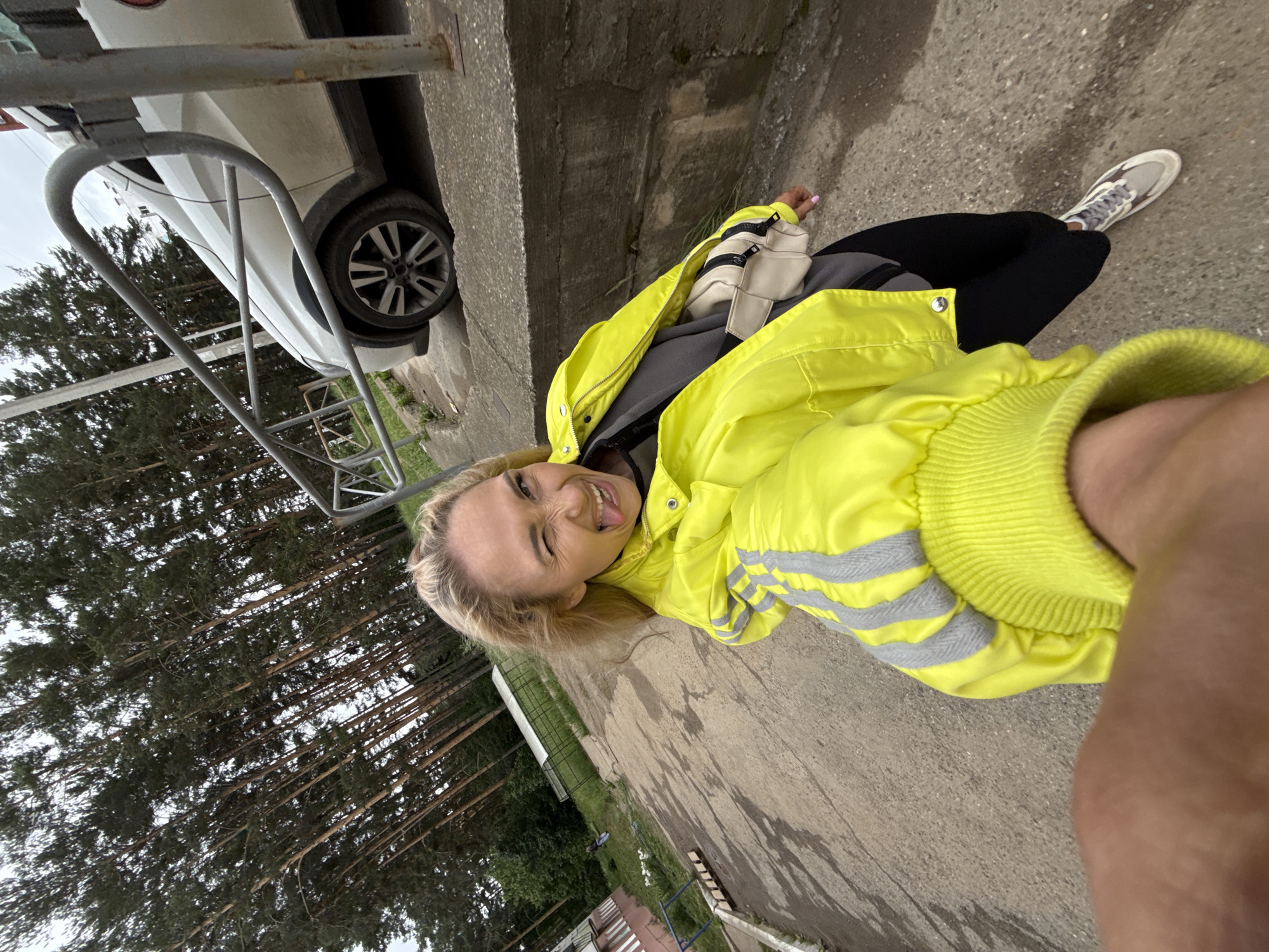 A wide-angle selfie taken on a concrete path in Chaikovsky, Russia, captures a blonde female making a playful expression. The subject, positioned centrally, winks with her left eye and sticks out her tongue. She wears a bright neon yellow jacket with grey reflective stripes on the sleeves and matching ribbed cuffs. Underneath, a dark grey top is visible, and she wears black leggings and grey and white athletic sneakers. A light beige fanny pack with black zippers is worn across her body.
The image is captured from a low angle, with a tanned forearm and hand holding the camera visible in the bottom left foreground. The concrete path, which appears wet with puddles or damp patches, recedes into the background.
To the left of the path, a multi-level building with reddish-brown and lighter-colored sections is partially visible. Further back, a green field contains a long, low greenhouse-like structure and a small, distant figure. A dense forest of tall pine trees dominates the left background and extends across the upper portion of the frame under an overcast sky.
To the right, a white SUV or crossover vehicle, appearing somewhat dirty, is parked behind a grey metal barrier or fence. This barrier, composed of curved and straight bars, extends along the edge of the path. A concrete curb or retaining wall separates the path from the higher ground where the car is located. More pine trees are visible in the background behind the vehicle.