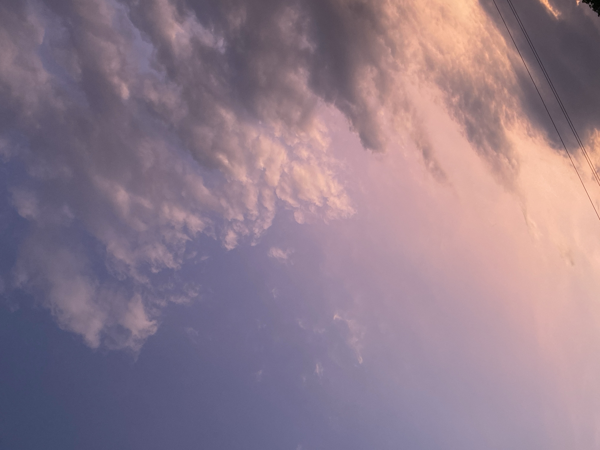 The image shows a sky with clouds, displaying a gradient of colors from purple to orange, possibly during a sunset or sunrise. There appear to be power lines visible in the upper right corner. There is no indication of Tinaquillo, Venezuela. No people, subjects, objects, actions, or interactions beyond the cloud formations are visible.