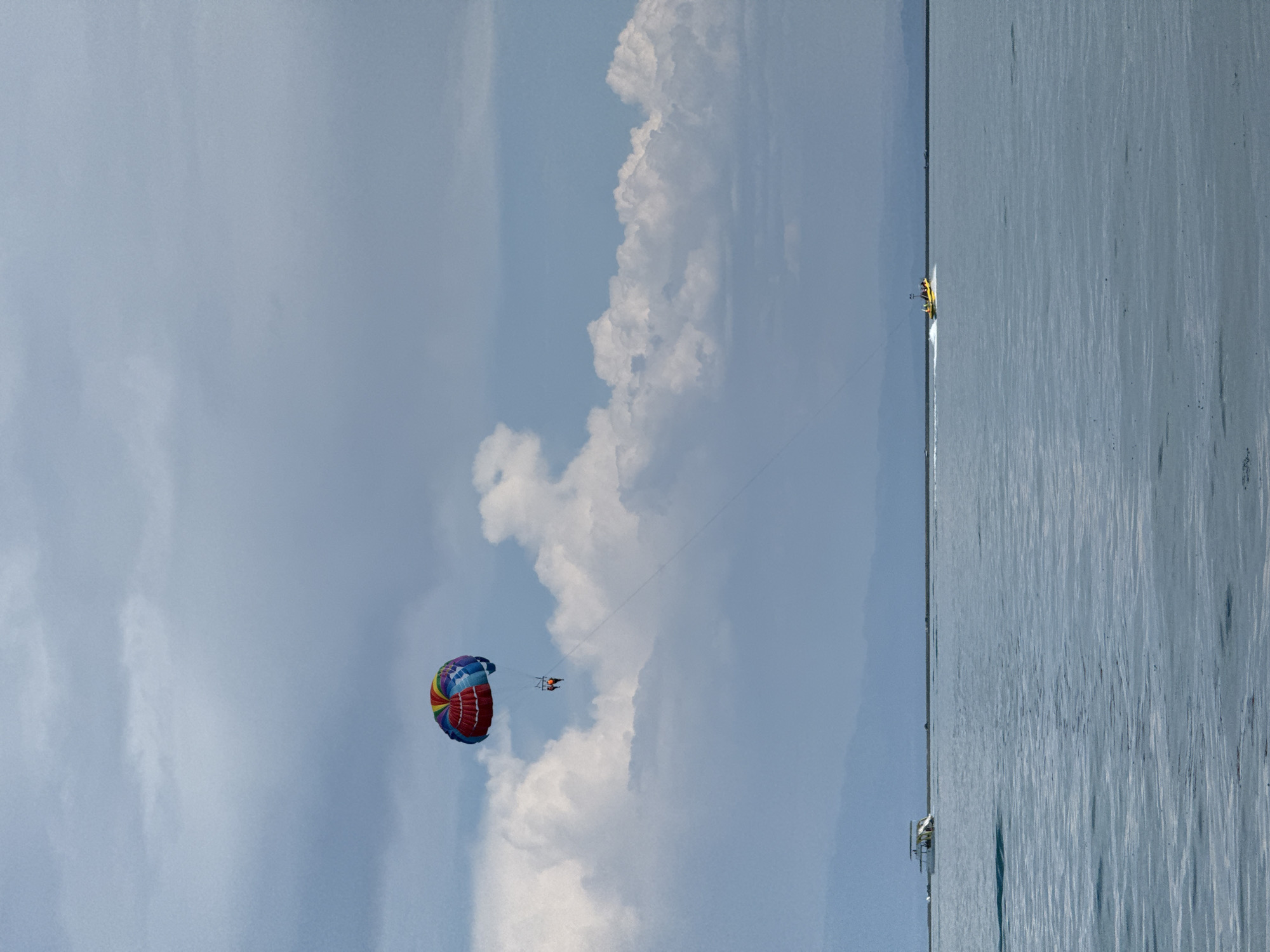 A wide shot captures a parasailer gliding through the sky over a calm, blue body of water. The parasail is colorful, with segments of red, yellow, green, blue, and purple. Two people are visible, suspended beneath the parasail. In the distance, a small white boat with a canopy is visible on the left side of the frame. Further to the right, a yellow speedboat is moving, creating a wake. The horizon line is low, with a faint, hazy mountainous landscape visible in the far distance. The sky is a muted blue, with scattered white clouds. The overall impression is of a recreational activity taking place in a natural setting. The location is identified as Akshy, Kazakhstan.