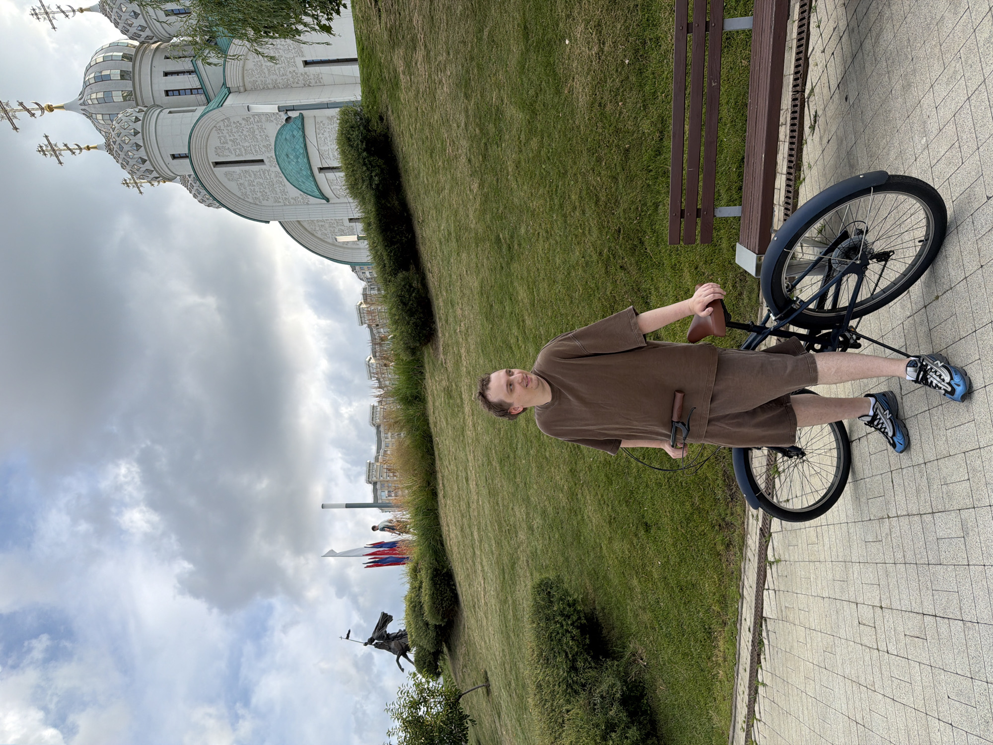 A young person stands next to a bicycle on a paved walkway. The person is wearing a brown t-shirt and shorts, and blue and black athletic shoes. They are holding the handlebars of the bicycle, which has a dark blue frame and fenders. To the left of the walkway is a grassy slope with a few bushes and trees. In the background, a large, white, ornate building with multiple domes and crosses is visible. The sky is cloudy, suggesting it might be overcast or a partly cloudy day. A brown wooden bench is also visible next to the walkway. The scene appears to be outdoors, possibly in a park or public area, with the distinctive architecture suggesting a religious building. The image is oriented sideways, with the ground appearing on the left and the sky on the right.