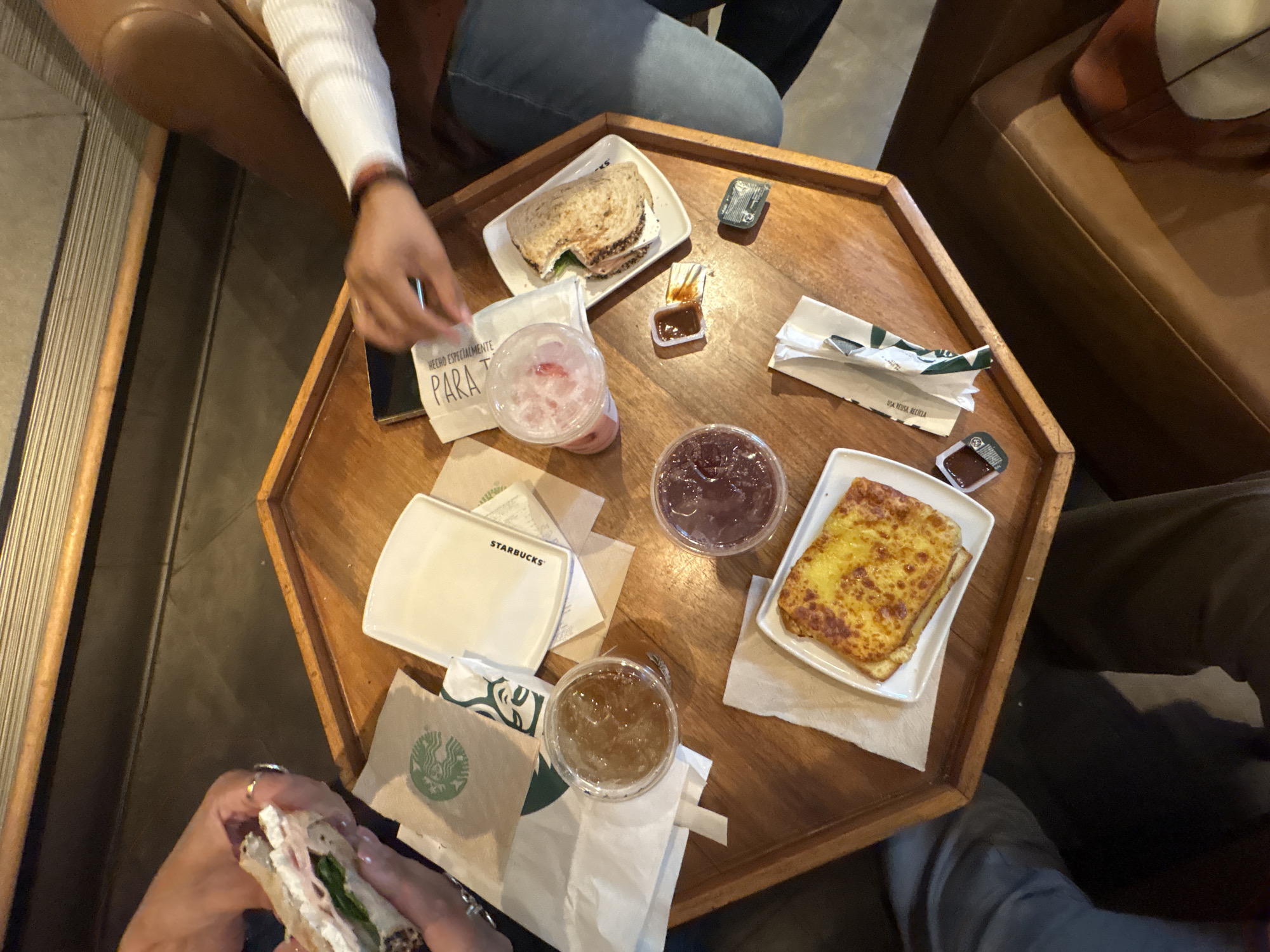 The image shows a wooden, octagonal table with several food items and drinks, likely at a Starbucks in Mexico City, Mexico. Two sandwiches are visible, one on the top left and another in the lower left corner, held by a person's hand with visible fingernails and a ring. A pink drink in a clear plastic cup is positioned near the top left sandwich. There is a dark, possibly jam-like substance in a clear plastic cup next to a square plate holding two slices of toasted bread or similar item. Two small sauce containers are on the table near the upper right. Napkins and Starbucks branded items are scattered around the table. Two people are partially visible in the image, seated around the table with their hands and legs shown. The setting appears to be indoors, possibly in a cafe or restaurant, with tan leather sofas partially visible in the background.