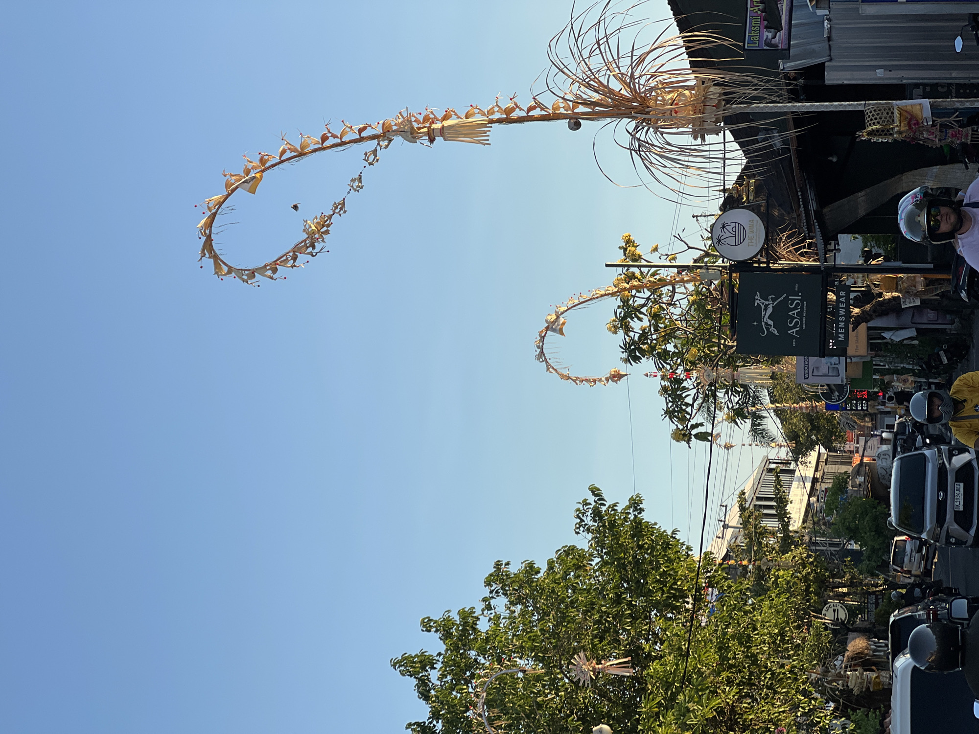In North Kuta, Indonesia, a street scene unfolds under a clear blue sky. Decorative, woven palm frond structures arch over the road, adorned with what appear to be small, dried flowers or fruits. Trees with dense green foliage line the left side of the street, some also decorated with similar woven ornaments. Buildings of varying sizes and styles are visible in the background, with shops and businesses indicated by signs. One sign reads "ASASI TAILORED MENSWEAR." Another sign displays "THE WIWA" with a palm tree graphic. Traffic is present, with several cars and motorcycles on the road. People are visible on the motorcycles, some wearing helmets. The lighting suggests it is either early morning or late afternoon.