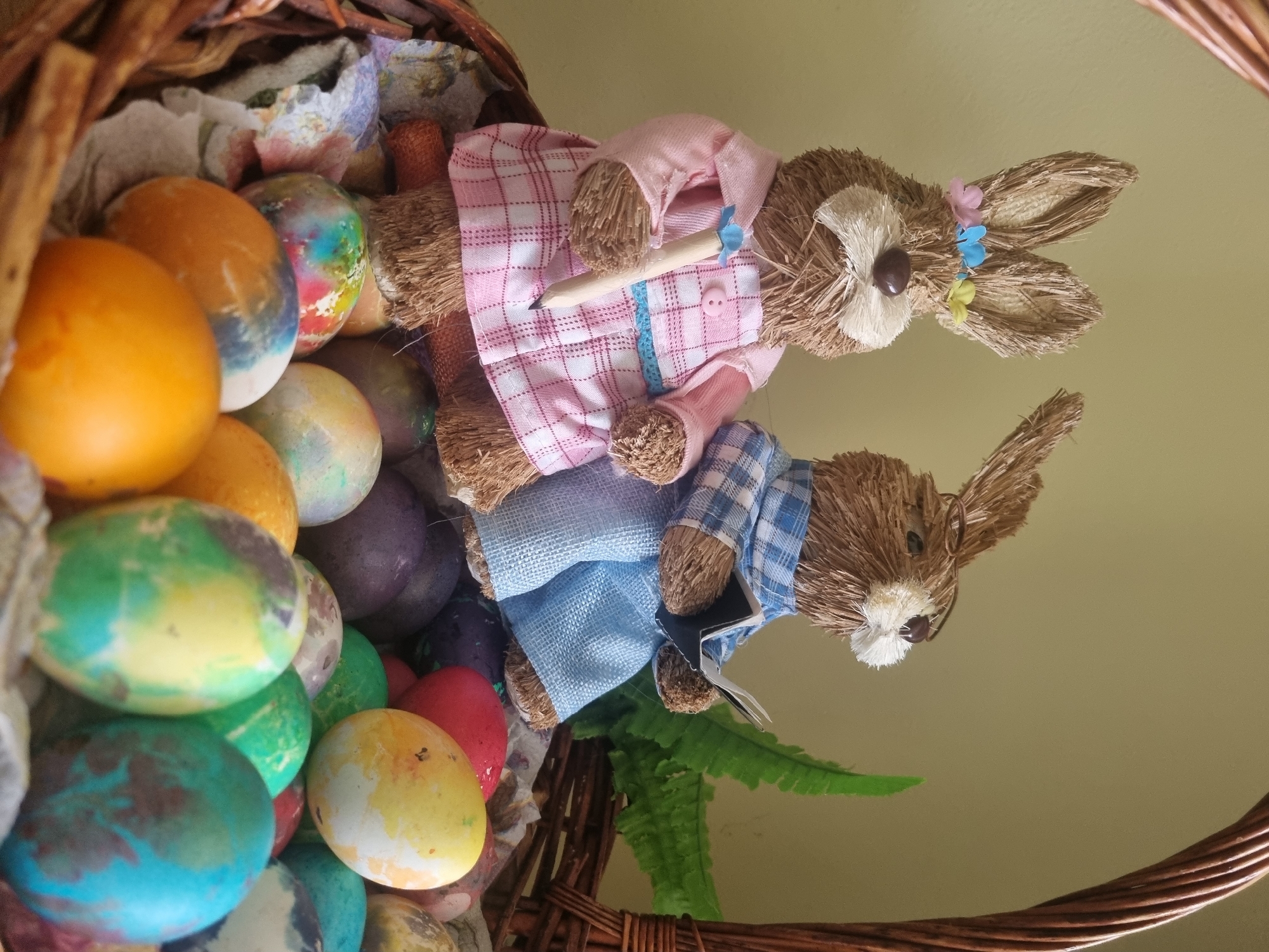A close-up shot displays an Easter basket filled with colorful Easter eggs and two straw rabbit dolls. The basket is made of woven brown material, and it is filled with a variety of Easter eggs painted in shades of orange, green, blue, yellow, red, and purple.
Two straw rabbit dolls are positioned on top of the Easter eggs. The top rabbit is wearing a pink and white checkered dress, and it has a pencil held in its paws. The bottom rabbit is wearing a blue and white checkered shirt. Both rabbits have brown straw bodies and white straw faces.
A green fern leaf is tucked next to the bottom rabbit. The background is a plain light yellow wall. The location and city are unknown.