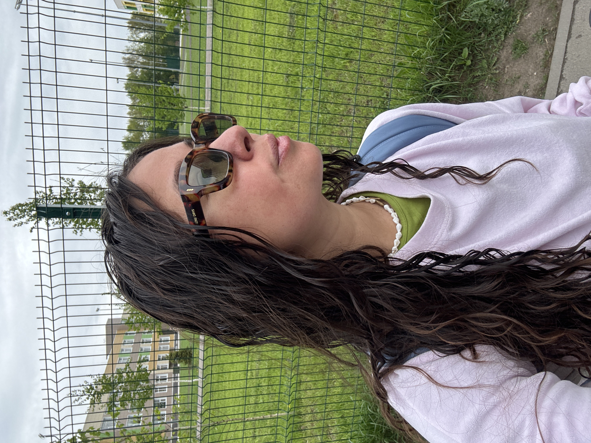 A young woman with dark, wavy hair and sunglasses is captured in a medium, angled shot. She is wearing a light pink sweatshirt over a green shirt and a white beaded necklace. She is standing near a wire fence, with green grass and a building visible in the background. The location may be in Kazan, Russia.