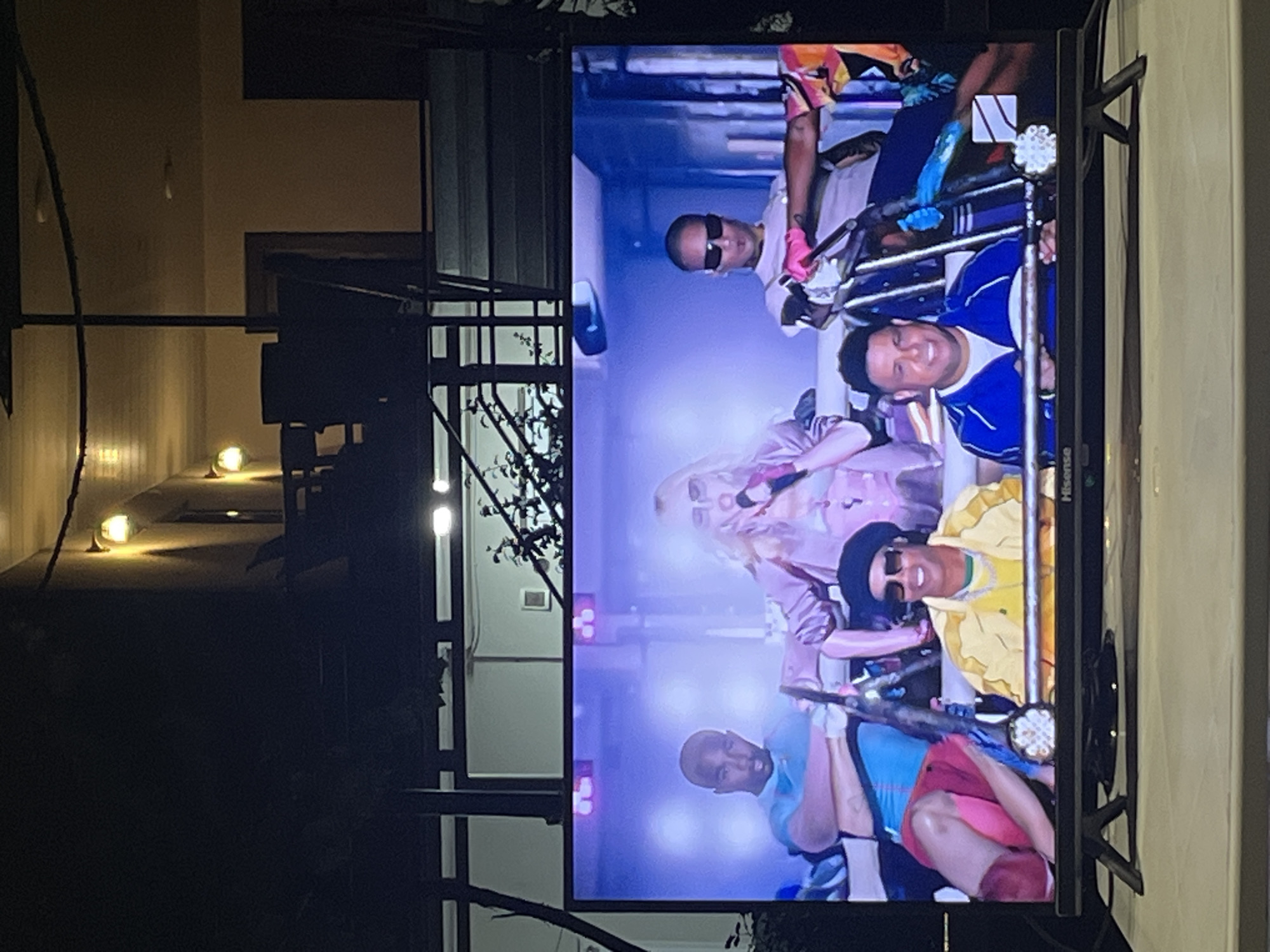 A television screen displays a scene from what appears to be a music video or performance. In the foreground of the screen, a woman with blonde hair and a pink outfit is singing into a microphone. To her left, a man in a blue shirt is seated, and to her right, a man in a light-colored shirt and sunglasses is also seated, with one leg extended. In the lower portion of the screen, two additional men are visible. One is wearing a yellow jacket and a black hat, and the other is wearing a blue jacket. The background on the screen is illuminated with purple and white lights, creating a vibrant atmosphere. The television set itself is a Hisense brand, with a black frame and visible support legs. The television is positioned on a light-colored surface, possibly a table or counter, in an outdoor setting at night. Above the television, a dark, blurred structure is visible, with outdoor lighting fixtures mounted on its ceiling. The overall impression is of an entertainment setup in a dimly lit outdoor space.