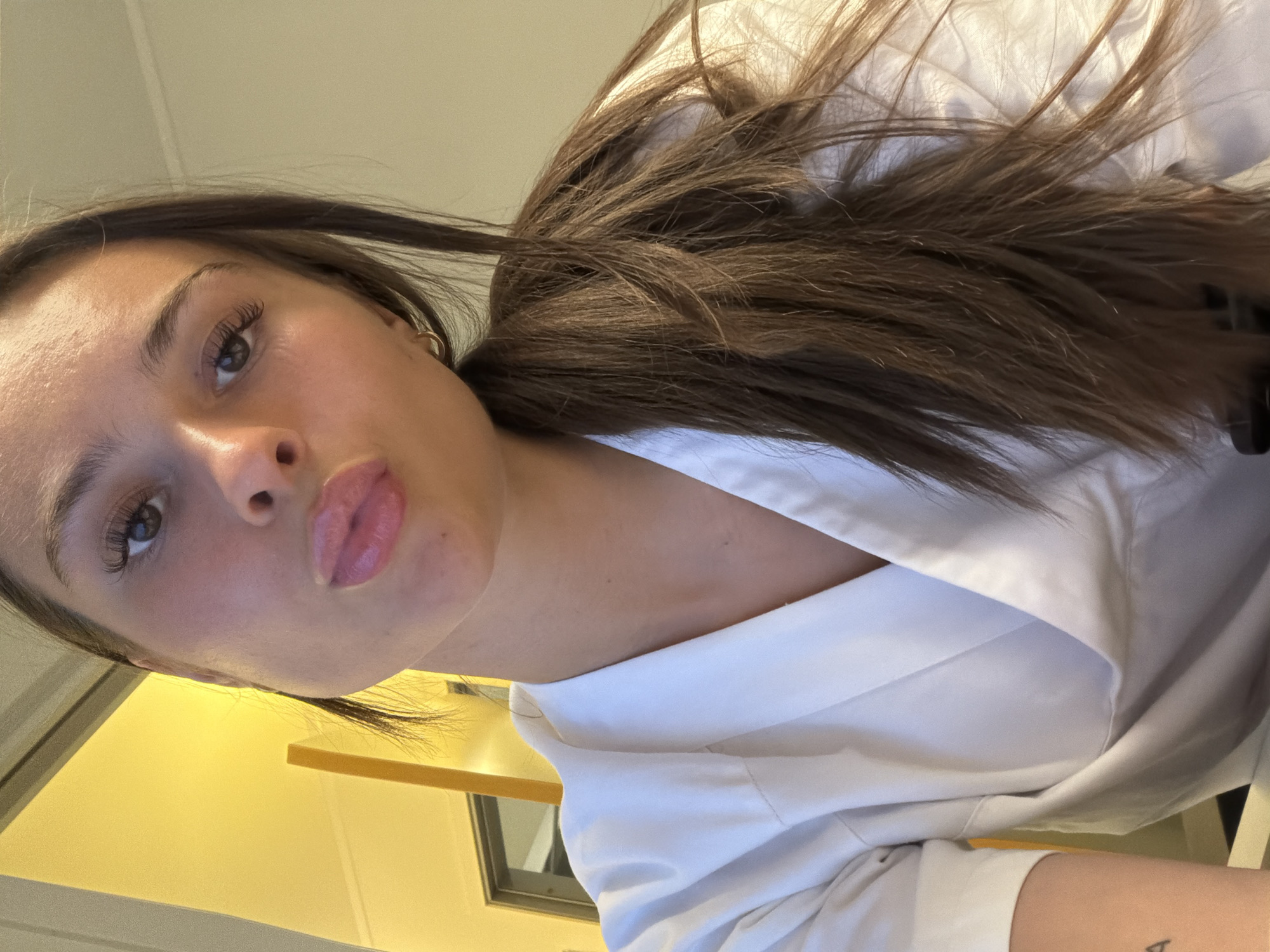 A young woman with long, brown hair pulled back into a ponytail is looking at the camera with her lips pursed. She is wearing a white V-neck top or uniform. Her right eye is visible, with long eyelashes. She has a small hoop earring in her right ear. The background is out of focus, but appears to be an indoor setting with a yellow wall on the left and a light-colored wall on the right. The image is a close-up selfie. This image does not contain information about the location in Harstad, Norway.