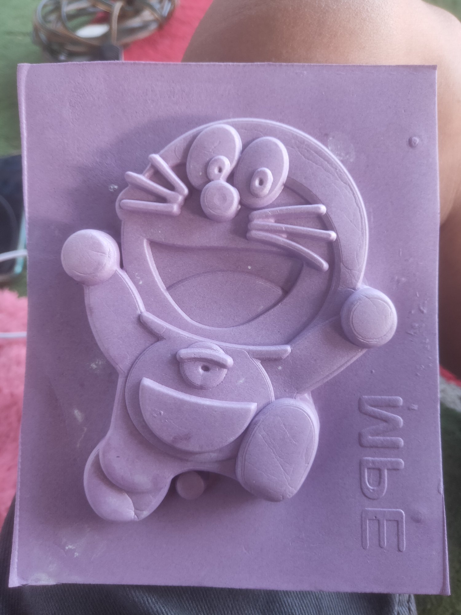 A close-up, top-down, outdoor shot shows a person holding a purple stamp of Doraemon in Serut, Indonesia. The stamp is in relief, with Doraemon's features raised from the surface. The background is blurred, with hints of greenery and possibly a pink surface. The lighting suggests an overcast day or shade. The person's skin tone is visible, indicating they are holding the stamp from behind. The stamp is rectangular, with the character filling most of the space. The text "IMBE" is embossed on the right side of the stamp. The overall color palette is dominated by purple and skin tones, with subtle green and pink accents in the background.
