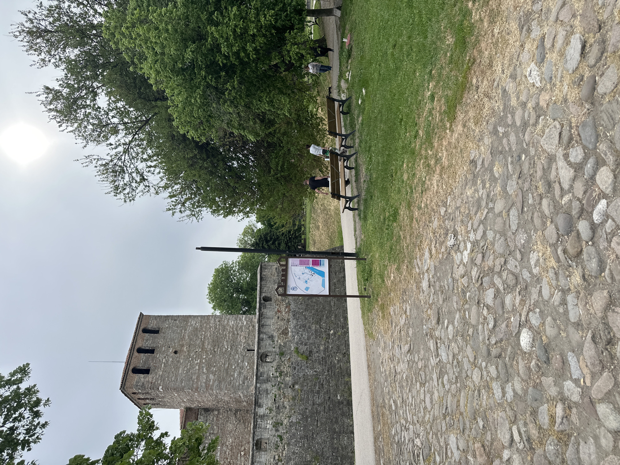 The image captures a scene in Vidin, Bulgaria. A stone tower, part of the Baba Vida Fortress, stands to the left. A map sign is positioned in front of the tower. Adjacent to the tower is a cobblestone path bordered by grass. Further into the scene, a row of benches is situated in a grassy area under a large tree, where several people are sitting or standing. The sky is overcast, and the sun is faintly visible.