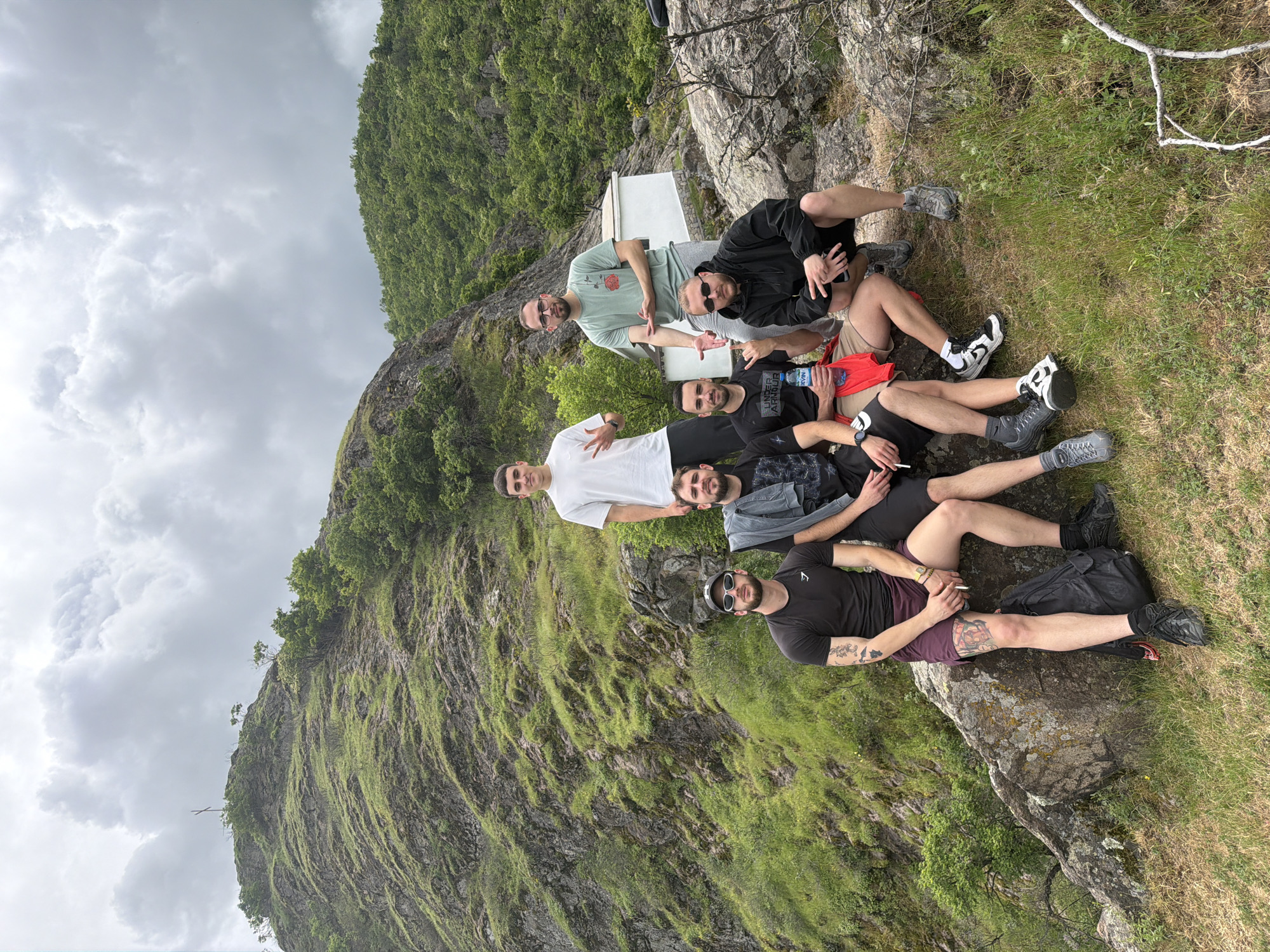 A group of seven men are positioned in a line, seated on a rocky outcrop near Ustina, Bulgaria. The background features a mountain covered in green vegetation and a cloudy sky. The first man is wearing a light green t-shirt and sunglasses. The second man is wearing a black hooded jacket. The third man is wearing a white t-shirt. The fourth man is wearing a gray jacket over a black shirt. The fifth man is wearing a black shirt. The sixth man is holding a cigarette.