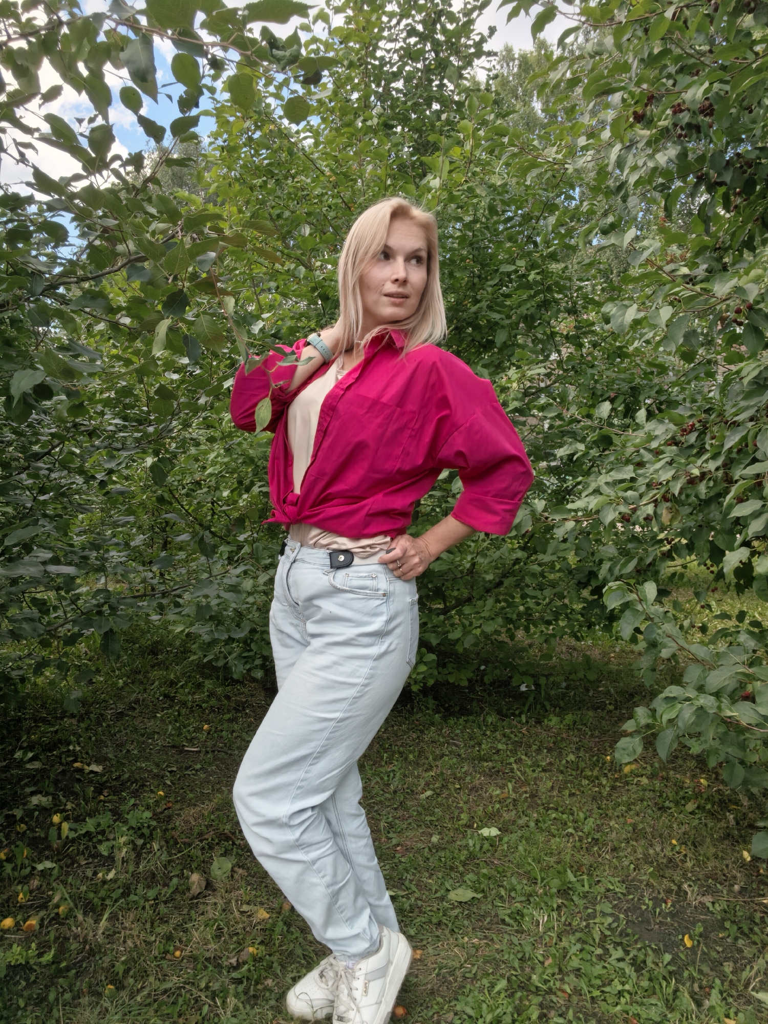 A fair-skinned woman with blonde hair, wearing a bright pink button-down shirt tied at the waist over a light beige top, light blue denim jeans, and white sneakers, stands outdoors in a leafy green environment. She is looking to her left, with her left hand on her neck and her right hand on her hip. The background consists of dense green foliage, likely trees and bushes, with some dark red berries visible on the branches. The ground is covered in grass and fallen leaves. The scene appears to be in a park or garden setting.