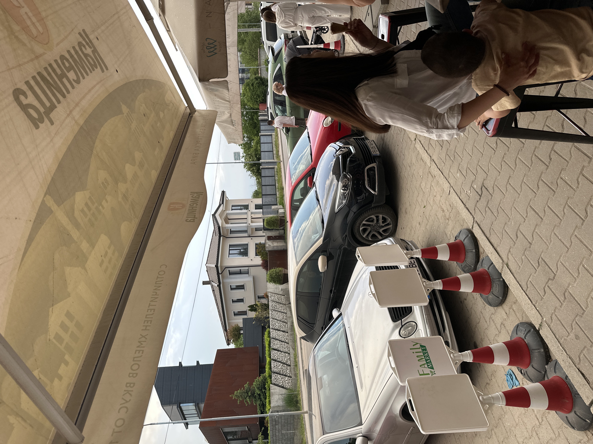 The image depicts an outdoor scene likely adjacent to a street. There are several cars parked. Prominent are a white car with "Family Market" on an advertising sign, followed by a black car and a red car. Street cones are present, positioned near the cars. In the background is a building with modern architecture. Part of a sunshade or awning with writing and an image is visible overhead. People are seated at a table on the right side of the image, including a woman in a white shirt with long dark hair. Based on the license plate format (PB xxxx PE) and language written on the awning (although mirrored), the location is likely Plovdiv, Bulgaria.
