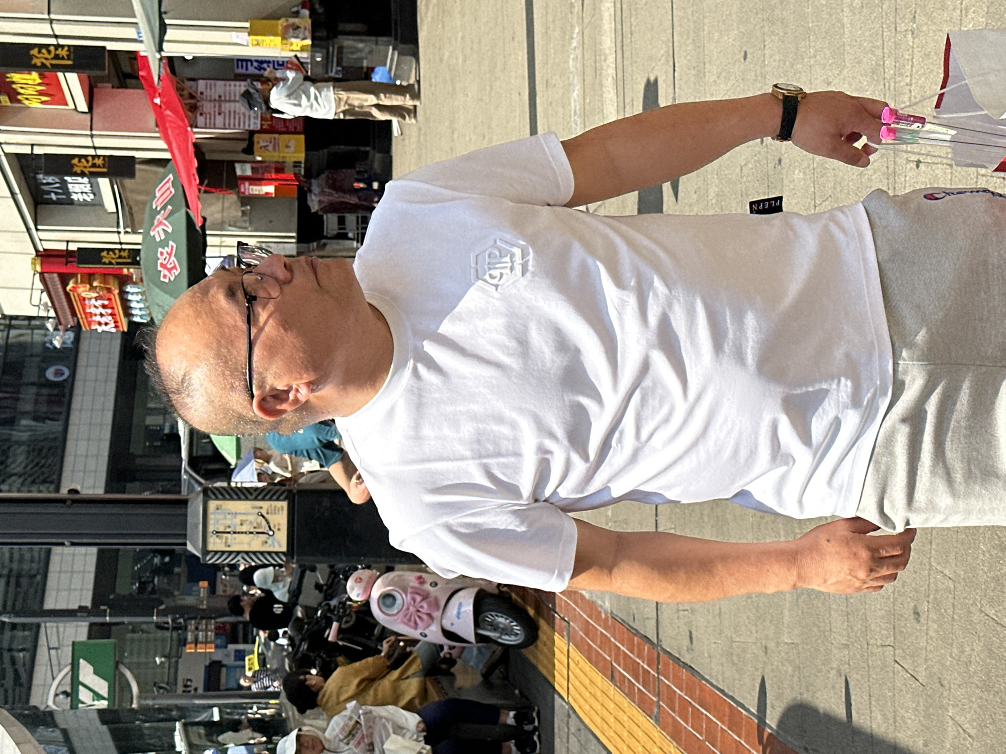 This media file captures an outdoor urban scene in Yuzhong District, China, under bright, direct sunlight. The main subject is a middle-aged man standing on a paved sidewalk, oriented towards the left side of the frame. He has short, dark hair and is wearing clear-framed glasses, a plain white short-sleeved t-shirt with a subtle, embossed hexagonal logo on the chest, and light grey sweatpants. A black tag with the text "PLEPN" is visible near his right hip. On his left wrist, he wears a gold watch with a black strap. In his right hand, he holds several transparent items with vibrant pink caps, possibly small umbrellas or decorative sticks.
The background reveals a bustling street or market area with numerous shopfronts and stalls. Multiple signs with Chinese characters are visible, including "十七路" (Seventeen Road) and other characters like "鲜" (Fresh), "佳" (Good/Excellent), and "口" (Mouth/Entrance), alongside red flags and green awnings. Several other people are present, including a vendor in a light-colored shirt and tan pants further down the street, and individuals seated near a pink child's scooter. A street sign displaying a map or route information is also discernible. The strong sunlight casts distinct shadows, suggesting it is midday. The man's expression is neutral as he looks slightly upwards.