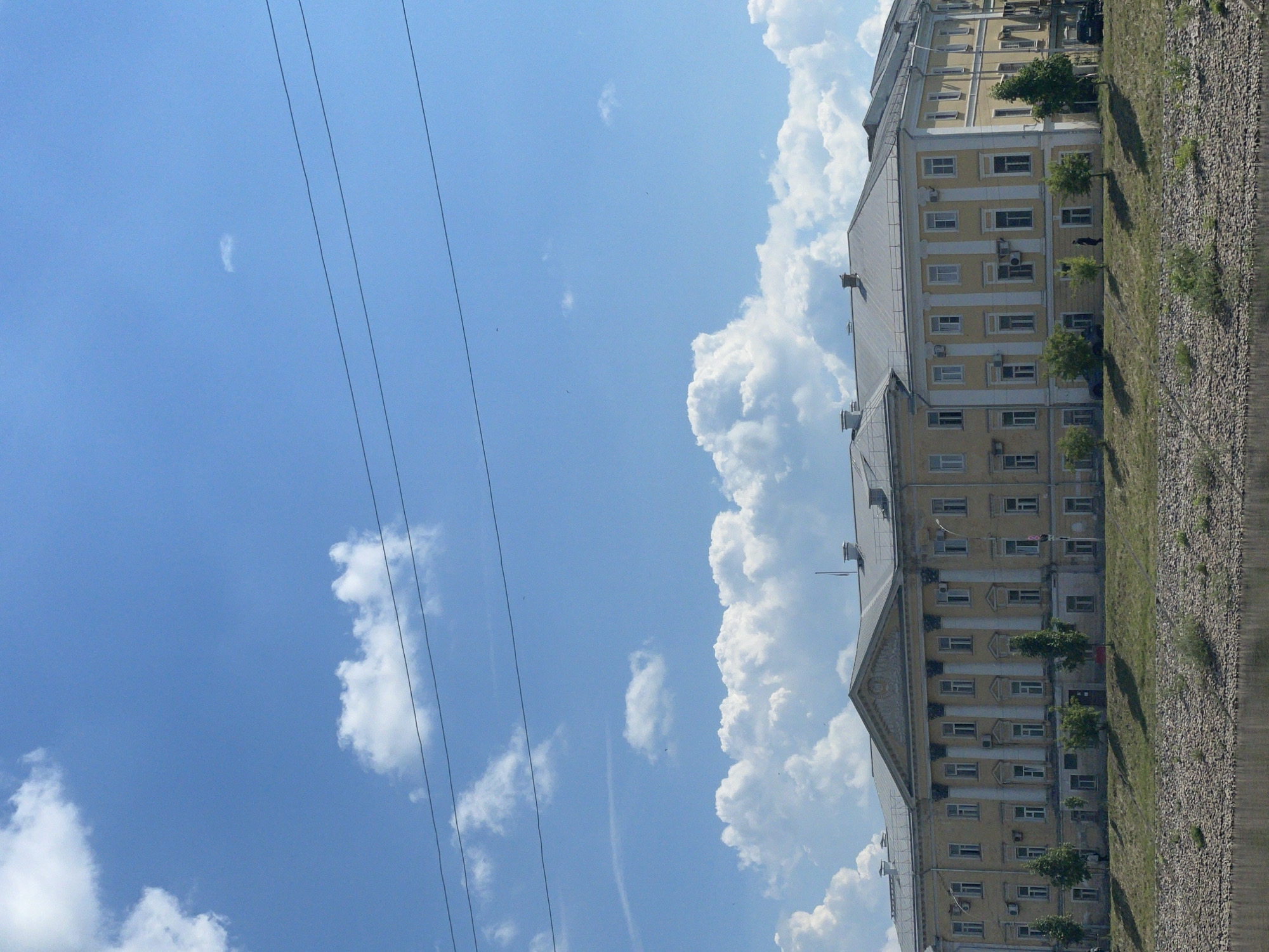 The image depicts a large, pale yellow, neoclassical building with a gray roof, located in Vologda, Russia. The building features a central pediment supported by columns, and rows of windows. In front of the building, there is a grassy area with several trees and a rocky embankment leading down to a body of water, likely the Vologda River, which runs along the foreground. The sky is blue with scattered white clouds. There are power lines stretching across the upper portion of the image. A few people are visible walking along the grass in front of the building, and a dark-colored car is parked on the right side of the image. The overall scene suggests a sunny day.