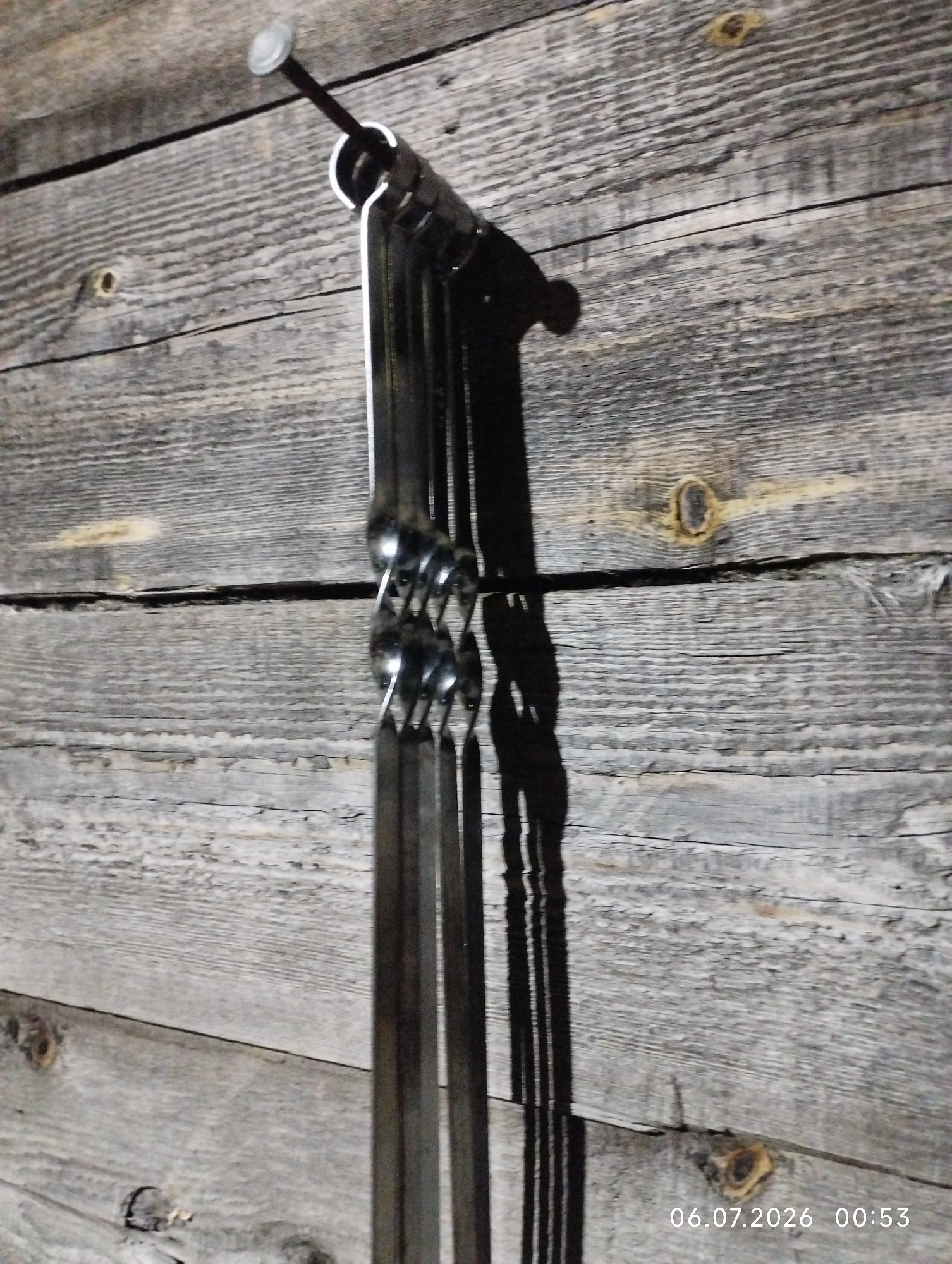 A close-up, slightly blurry, eye-level shot shows several metal skewers hanging from a large nail driven into a weathered wooden wall. The wooden wall is composed of horizontal planks with visible grain and knots, appearing aged and possibly outdoors. The skewers are dark metal and have a twisted or coiled section near what appears to be their tops. A shadow of the skewers and the nail is cast on the wall to the right. The bottom right corner of the image displays a timestamp: "06.07.2026 00:53". The location is identified as Pokosnoye, Russia.