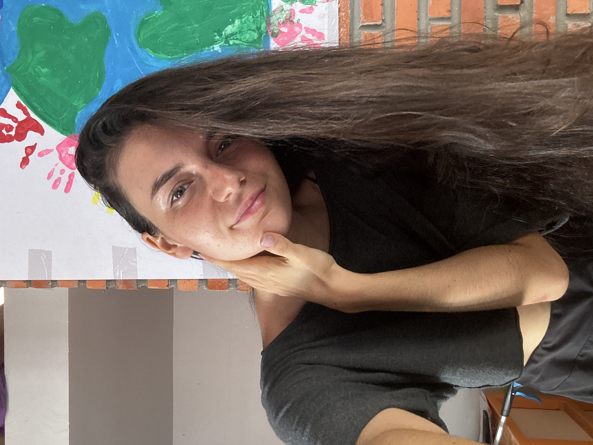 A woman with long brown hair, wearing a black shirt, poses with her hand under her chin in front of a painting of the Earth done with finger paints, and a red brick wall. The location is unspecified, and not clearly identified as Mũi Né, Vietnam.