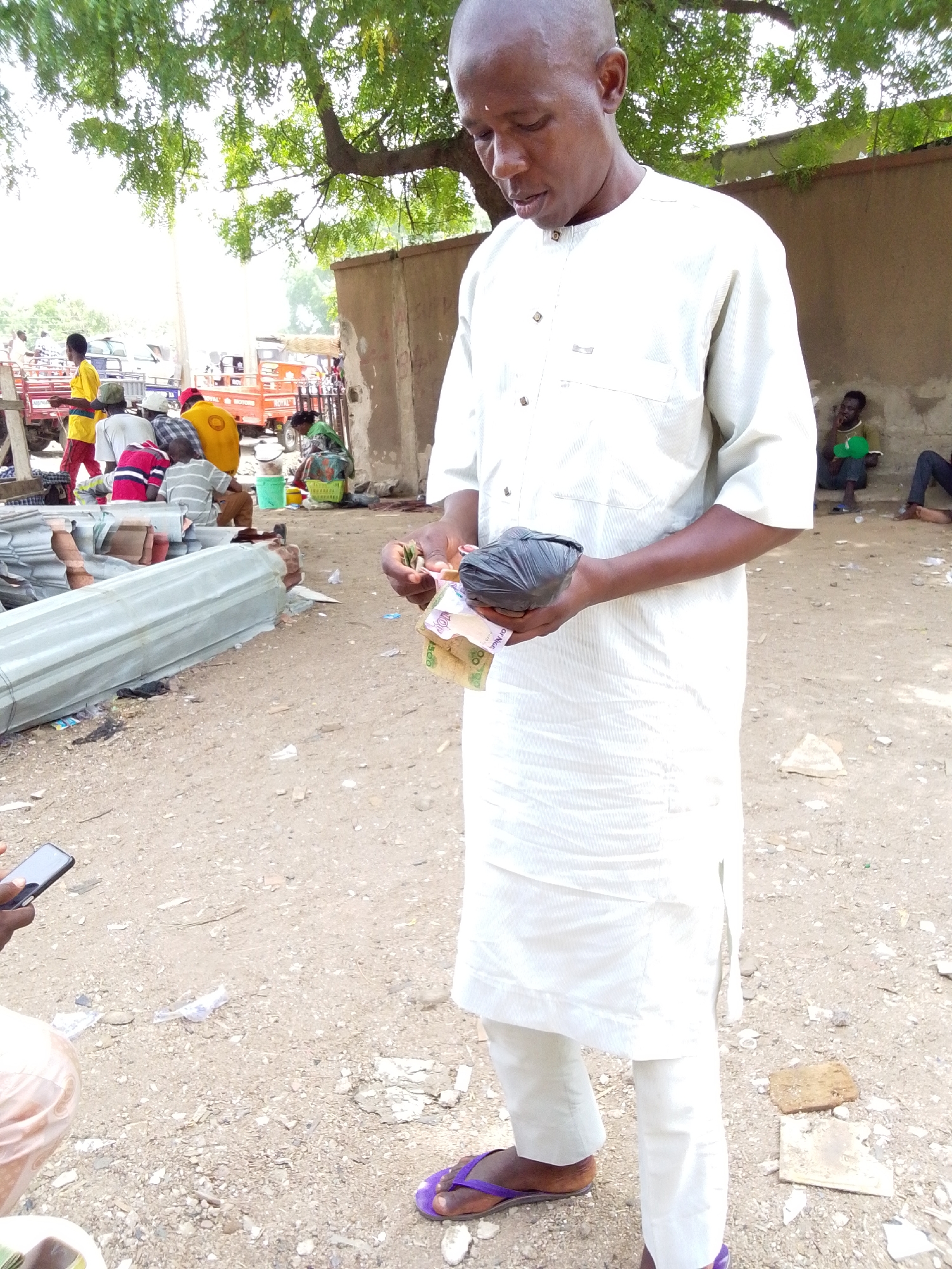 The photo shows a man standing in an open, outdoor area with a sandy, uneven ground. He is wearing a white tunic-like garment and white pants, and purple sandals. He's holding a black plastic bag in one hand and a small amount of Nigerian currency in the other. He appears to be looking at the money.
In the background, there are people and vehicles that indicate an active environment, possibly a market or roadside gathering. There's a green-leafed tree overhead. A building wall is visible to the right of the man. Based on the information provided, the location is Jimeta, Nigeria.