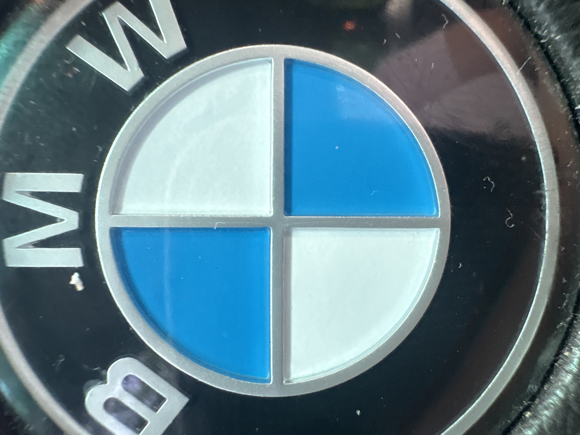 This media file features a close-up, high-resolution shot of a BMW logo. The logo is circular, prominently displaying the letters "BMW" in a metallic, silver color against a black outer ring. The inner circle of the logo is divided into four quadrants by a silver cross. The top-left and bottom-right quadrants are white, while the top-right and bottom-left quadrants are a vibrant blue.
Notable details include signs of wear and dust accumulation on the logo. Small white specks of dust are visible on the black outer ring, and some lighter scuff marks or reflections are present on the black surface. Similar dust particles and minor imperfections can be seen on the blue and white segments of the inner circle. The silver trim separating the quadrants and forming the outer edge of the inner circle also shows some subtle wear. The lighting suggests an indoor setting or diffuse light, with some reflections on the metallic and colored surfaces. The visible text is "BMW". The overall impression is an authentic, used BMW emblem, possibly from a vehicle or merchandise.