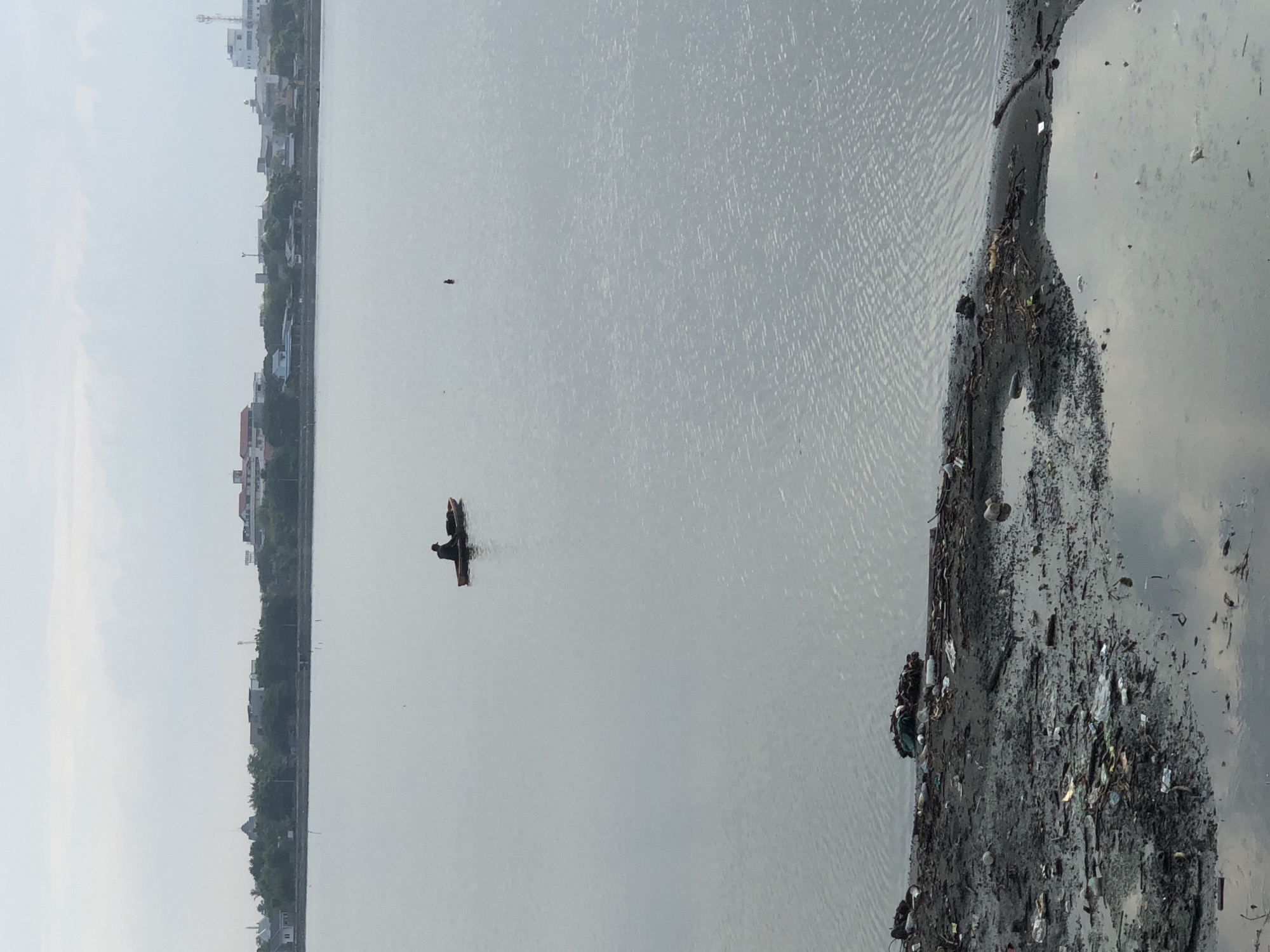 A person is visible in a small boat on a large body of water, possibly a river or bay, in Lhokseumawe, Indonesia. The water is calm with some ripples reflecting the overcast sky. On the distant shore, a line of trees separates the water from a cityscape with buildings of varying sizes and a prominent cell tower. The foreground of the image shows a muddy bank littered with debris, including plastic bottles, wrappers, and other refuse. The overall scene depicts a mixture of natural elements and urban development, with a stark contrast between the somewhat distant, developed shoreline and the polluted foreground.