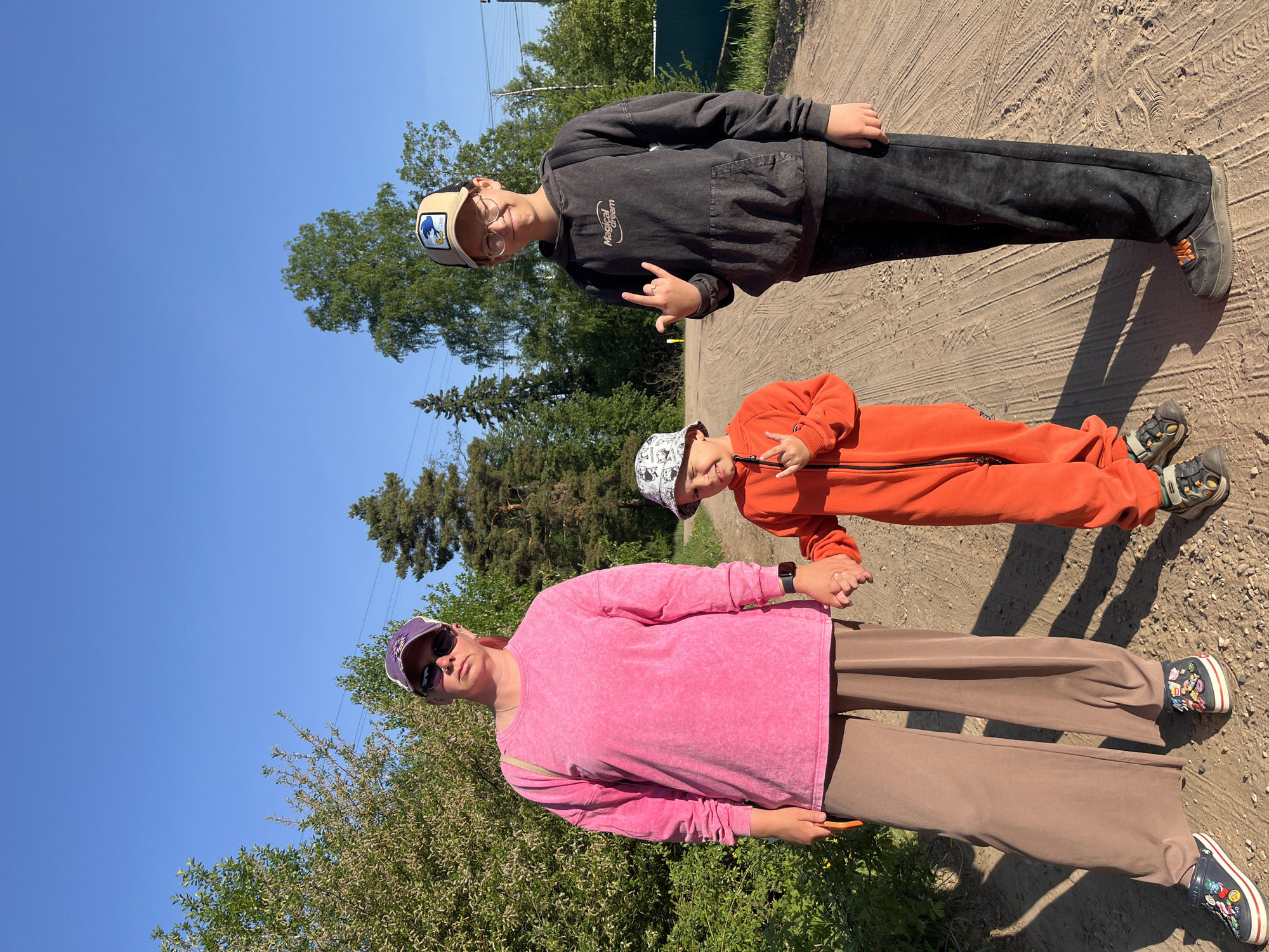 A medium shot depicts three individuals standing on a dirt road under a bright, clear sky. Tall trees and greenery line the sides of the road. At the top of the frame, an older boy with glasses wears a black hoodie, dark pants with sparkles, and a baseball cap. He makes a hand gesture. Below him, a younger child in an orange jumpsuit and patterned bucket hat mimics the hand gesture. At the bottom of the frame, an adult wearing a pink top, wide brown pants, sunglasses, and a purple hat holds the younger child's hand. The location may be in or near the city of Chaikovsky, Russia.