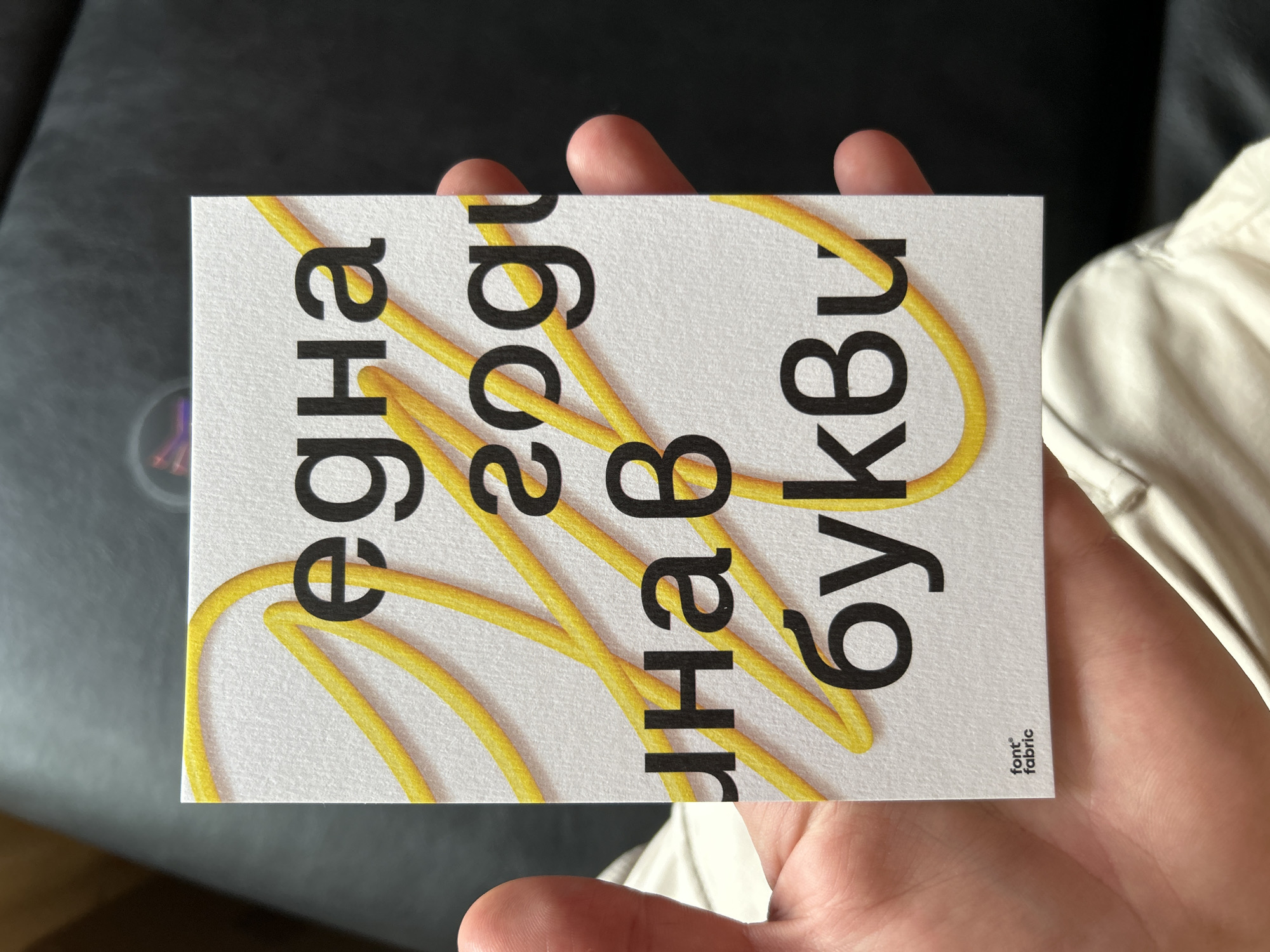A hand holds a rectangular card with Cyrillic text overlaid with yellow, noodle-like illustrations. The text, printed in large black font, reads "една году нав букви". In the lower right corner, in a much smaller font, is the text "font° fabric". The background is blurred and shows a dark surface (possibly leather) and light-colored fabric. The location is not discernible from the image.