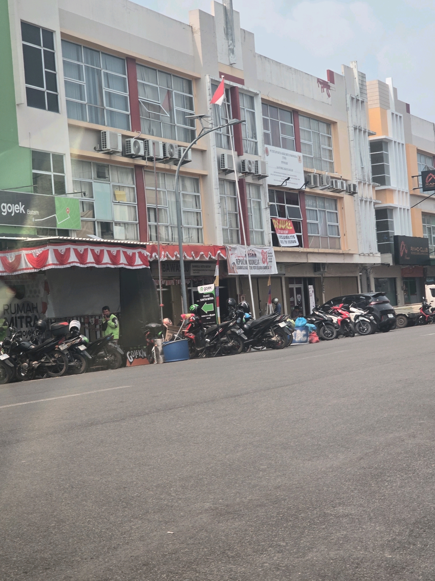 The image shows a street scene in Batam, Indonesia. In the background, a multi-story building with large windows and air conditioning units is visible. There are several businesses on the ground floor, including one with a red and white awning and signs that say "RUMAH MITRA" and "Gojek Batam". A man in a bright green jacket is standing near a parked motorcycle. Several other motorcycles are parked along the street. The street itself is paved and appears to be a busy area. The sky is overcast.