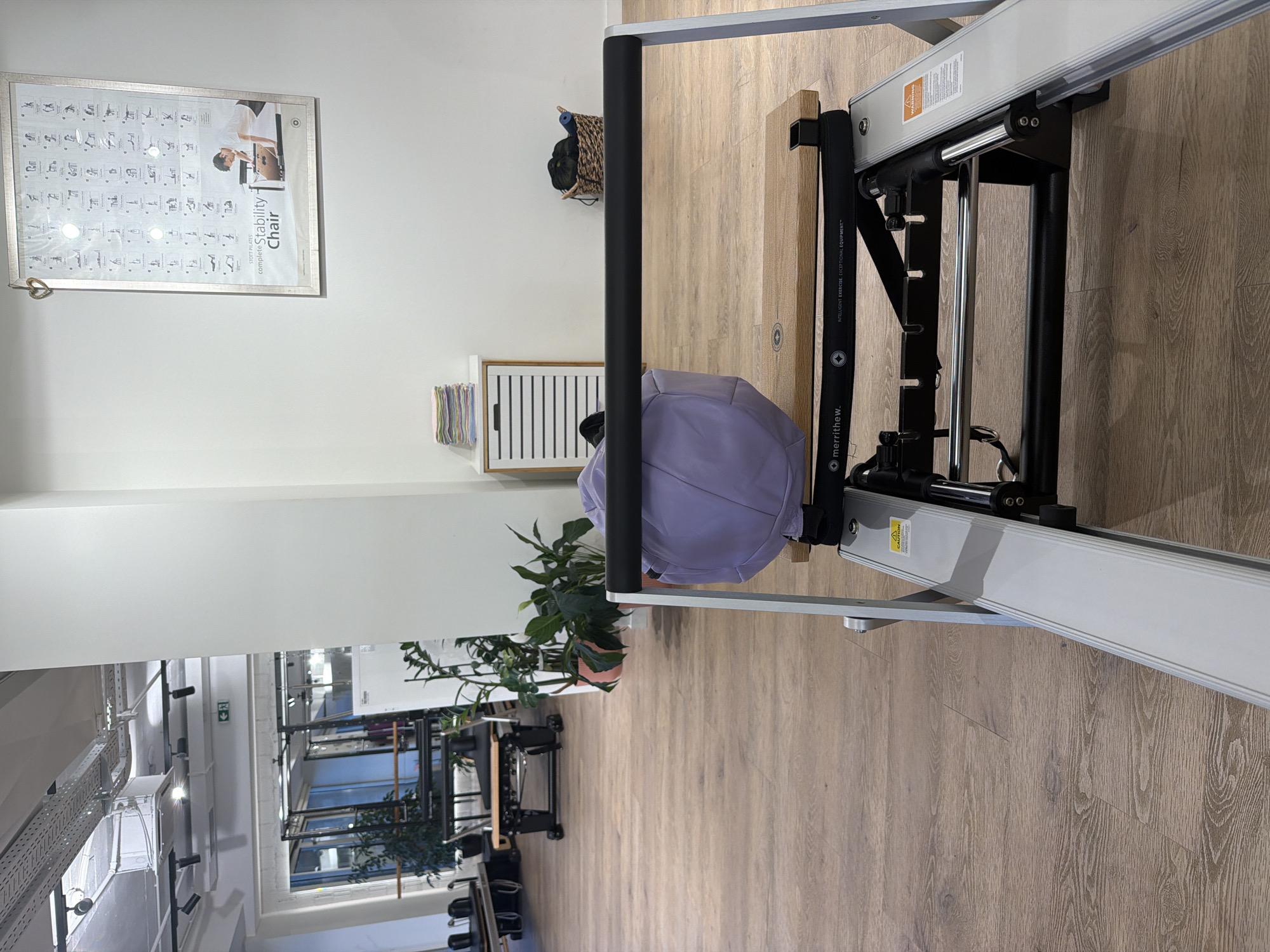 The image shows the interior of a brightly lit fitness studio, possibly a Pilates studio, with a wooden floor. In the foreground, a piece of Pilates equipment, likely a reformer, is visible. A light purple duffel bag rests on the equipment's footplate. In the background, there are other pieces of fitness equipment, including reformers and what appears to be a ballet barre. Several potted plants are strategically placed around the room. On the right wall, a framed poster displays various exercises, identified as "STOTT PILATES complete Stability Chair." To the left of the poster, a small white cabinet with horizontal drawers holds a stack of folded colorful towels. Further to the right, a woven basket contains what appear to be workout accessories. The environment suggests a clean, organized, and professional space for physical training. There are no people visible in the image. The location is stated to be Boreti, Montenegro.