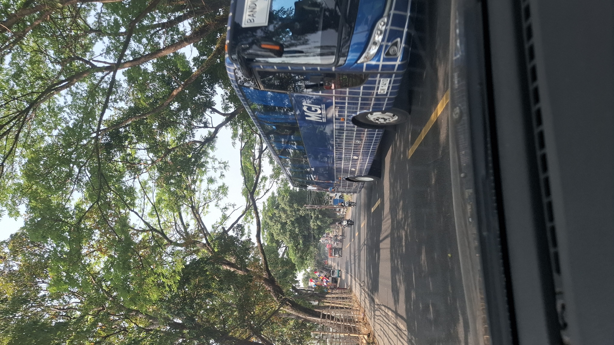 A bright, daytime, street-level shot captures a blue bus with a white grid pattern on its side traveling down a paved road lined with trees. The bus has "MGI" written on its side in white lettering. The road is marked with yellow lines. In the background, other vehicles, including motorcycles and cars, are visible, along with more trees and some buildings. The image appears to have been taken from inside another vehicle, as the dashboard is visible in the foreground. The overall scene suggests a busy street in Cikaroya, Indonesia, possibly during the day given the bright lighting and shadows cast by the trees.