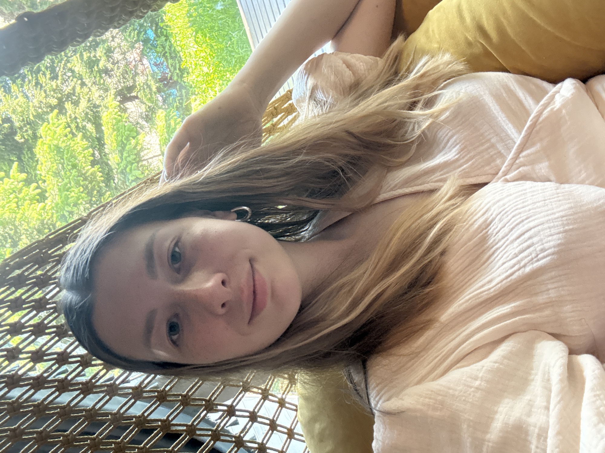 A young woman with long blonde hair lies in a hammock. She is wearing a light pink shirt. The background shows green foliage, possibly trees. The hammock is constructed of woven rope. A yellow pillow is behind her. The location is unknown.