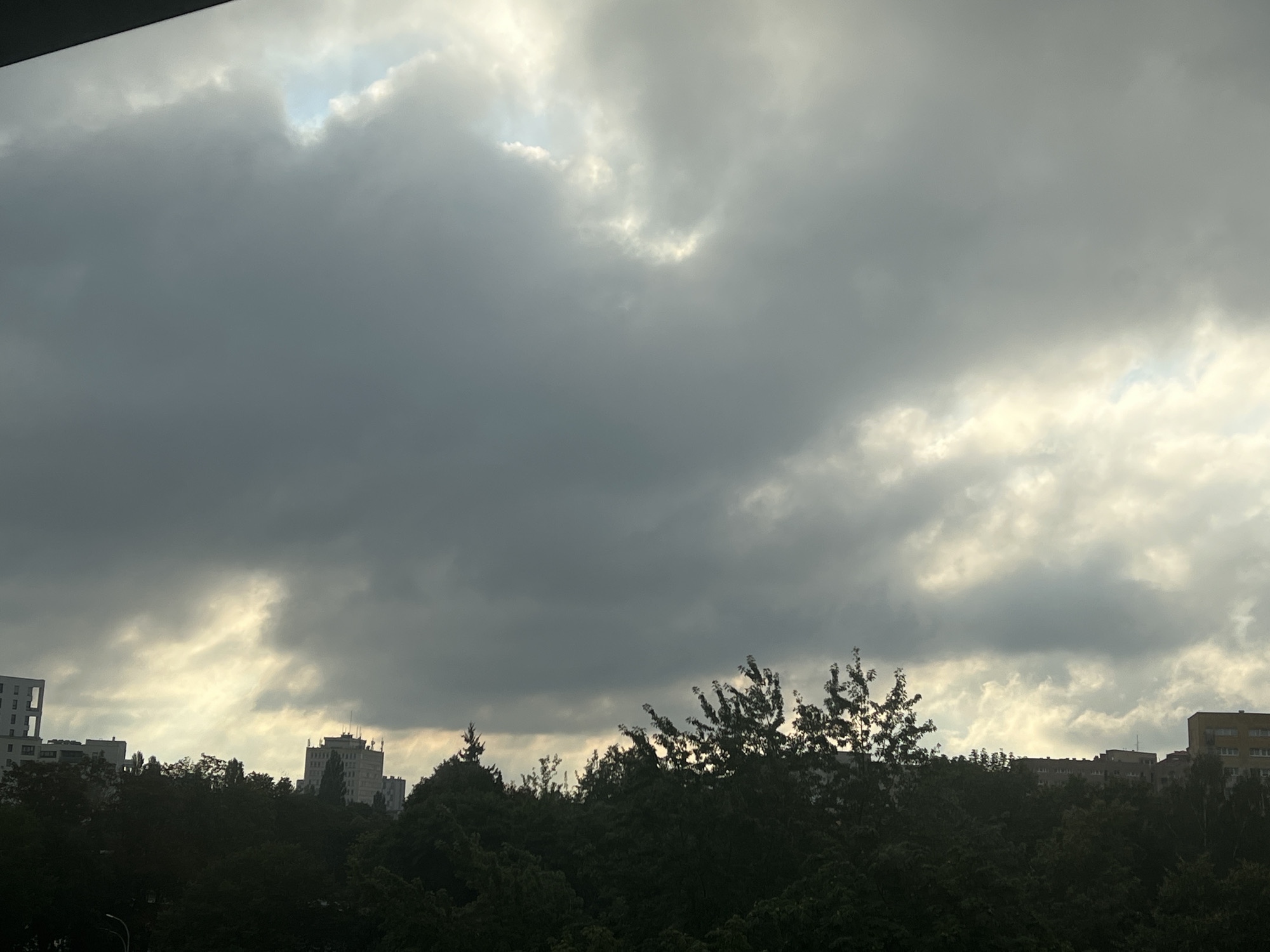 The image captures a view of the sky and rooftops in Warsaw, Poland, likely from an elevated vantage point. The sky is dominated by dramatic, dark clouds with patches of bright, illuminated sky peeking through, suggesting a transition in weather or the setting of the sun. Silhouetted against the sky are the tops of dense trees that form a dark green band across the lower portion of the image. Above the trees, the upper floors of several multi-story buildings are visible, including a modern, tall white building on the far left, a lighter colored building with a more traditional facade in the middle, and a yellow building with many windows on the far right. The overall impression is one of an urban landscape under a brooding sky.