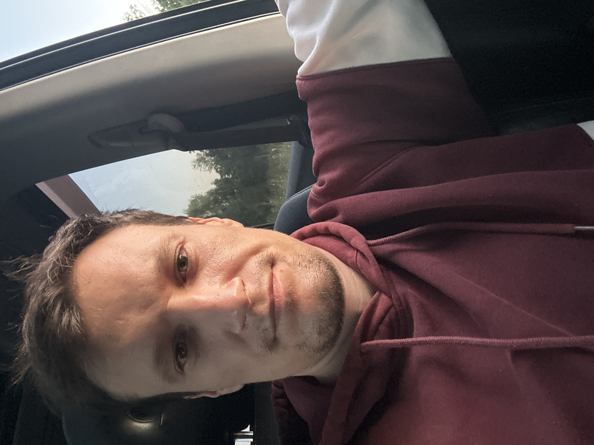 A man is seated inside a vehicle, his upper body filling the frame. He is looking directly at the camera with a neutral expression. He has short, dark brown hair and stubble on his chin. He is wearing a maroon hooded sweatshirt with white accents on the sleeves. The background suggests an outdoor environment with trees visible through the car window, consistent with a location in or near Makiv, Ukraine. The lighting indicates it is daytime, possibly late afternoon or early evening.