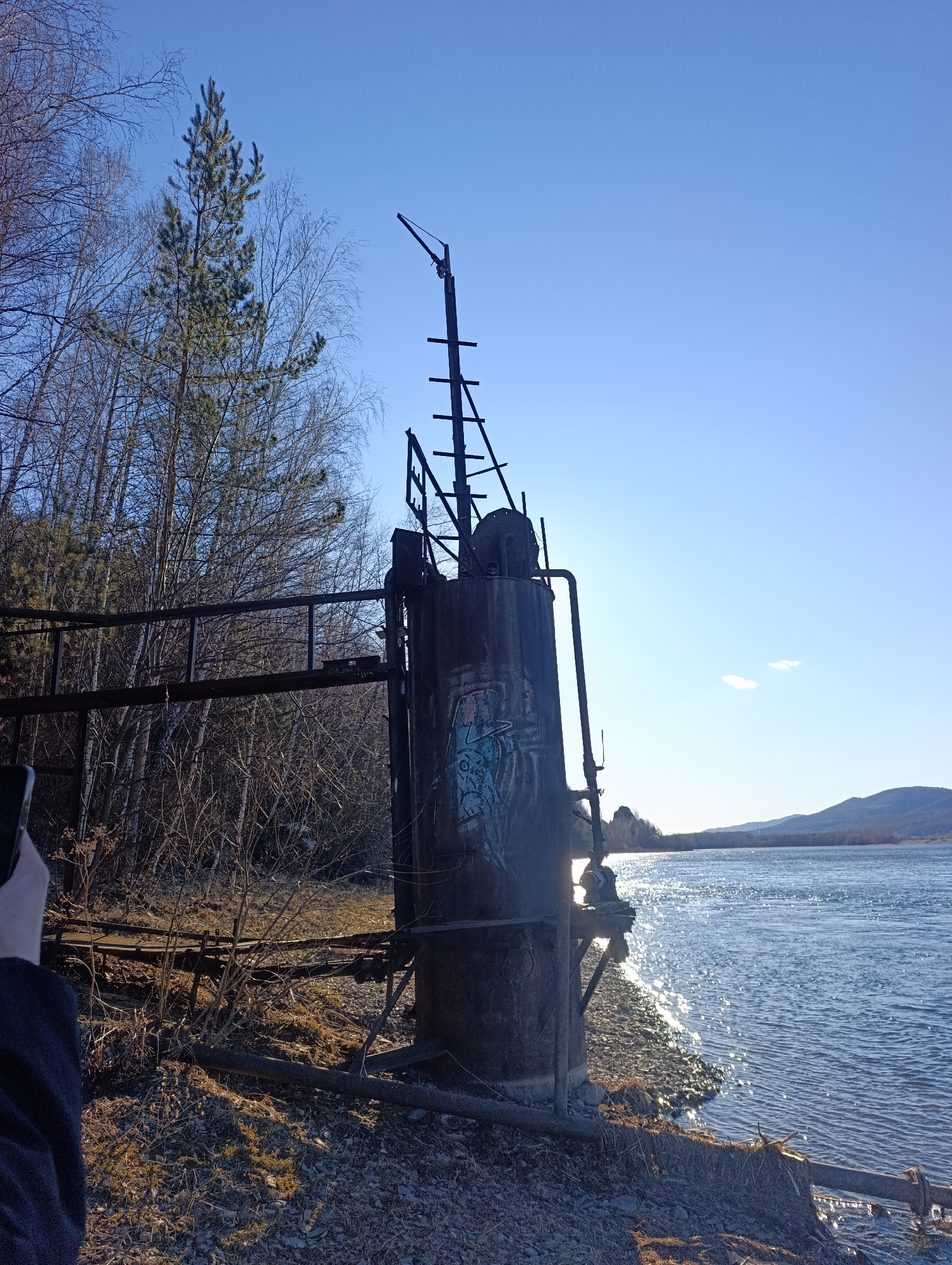A rusted, cylindrical metal structure with attached pipes and a skeletal metal framework stands on a pebble-strewn shore next to a body of water, likely the Yenisey River. Graffiti marks the side of the cylinder. A metal walkway extends from the structure into a thicket of bare trees. In the background, a mountain range is visible under a clear blue sky. The location is likely in the vicinity of Krasnoyarsk, Russia, given the geography. A person's arm is partially visible in the bottom left corner of the frame, holding a device, presumably a phone or camera. The sun is reflecting off the water's surface.