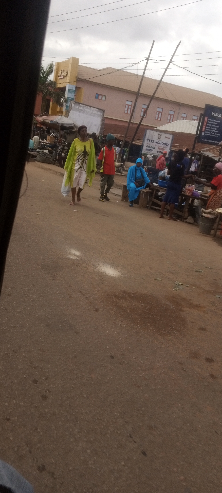 The image captures a street scene in Sabon Tasha, Nigeria. Several people are visible, including a woman in a light green top and white shorts, walking towards the left. Next to her is a person in a red shirt and green hat. Further to the right, a person in a blue garment is seated. Several other people are standing or sitting around makeshift stalls and benches.
In the background, buildings are visible, including one with "VYTE SCHOOLS" and "VINCO" signs. Also "ST. STEPHEN'S CHURCH" is visible. Utility poles with wires are prominent in the sky. The road surface is uneven with some patches of debris.
