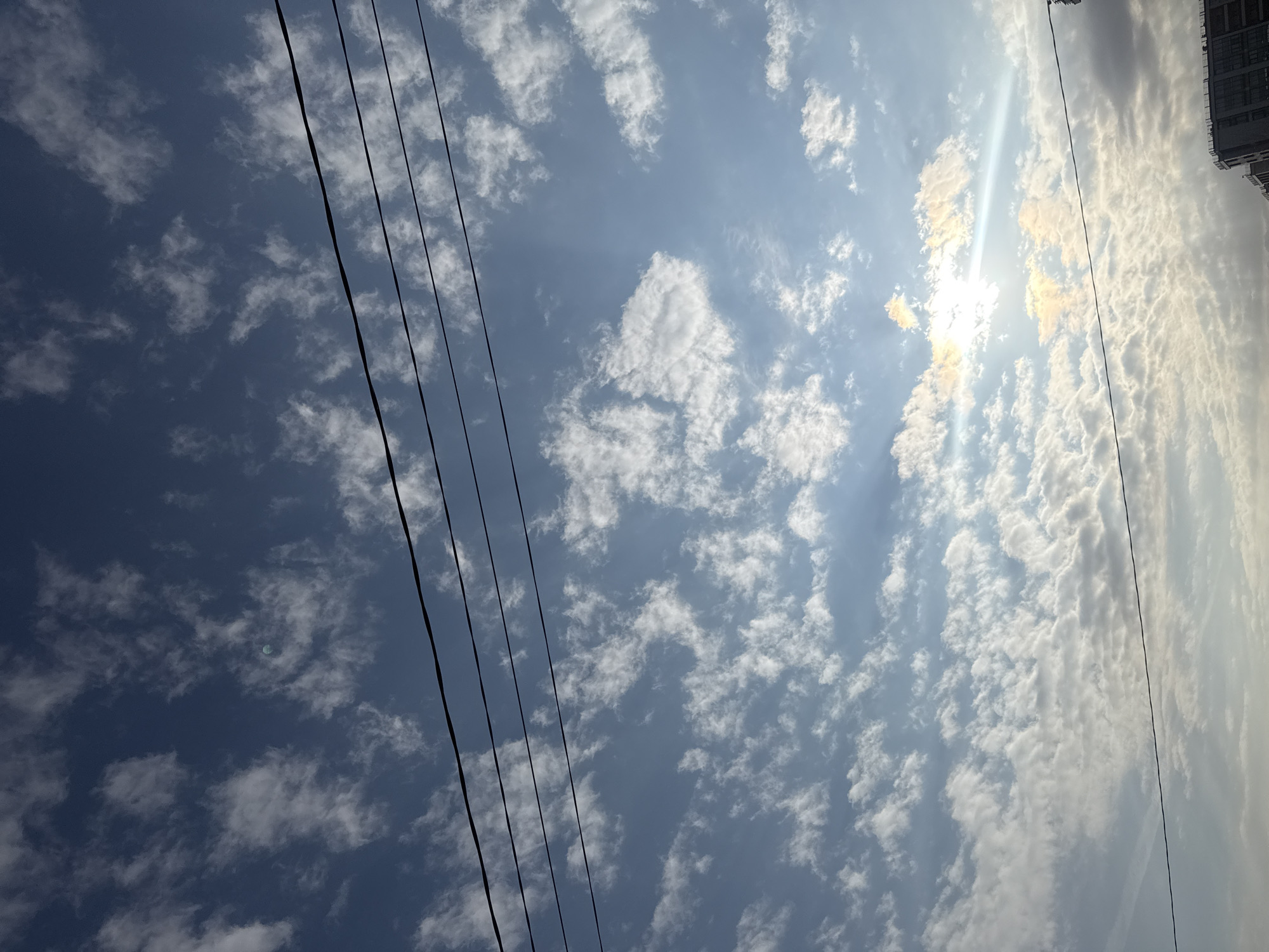 The image shows a partly cloudy sky with power lines running across the lower-left side. The sun is visible through the clouds on the right side of the image, creating sun rays. A building is partly visible in the upper-right corner. The location is likely Saint Petersburg, Russia.
