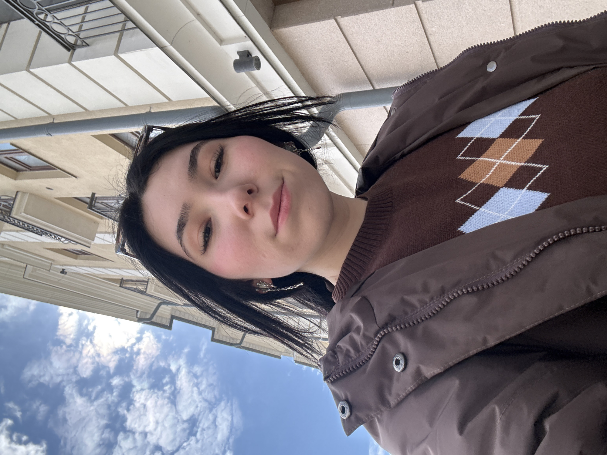 A young woman is posing for a selfie. She has dark hair, fair skin, and appears to be wearing a brown argyle sweater and a brown jacket. The background is a mix of blue sky with clouds and the facade of a light-colored building with architectural details. The image is taken at a low angle, looking up. While the exact location in Kazan cannot be determined with certainty from the image, the architectural style and general urban setting suggest it is likely a city in Russia.