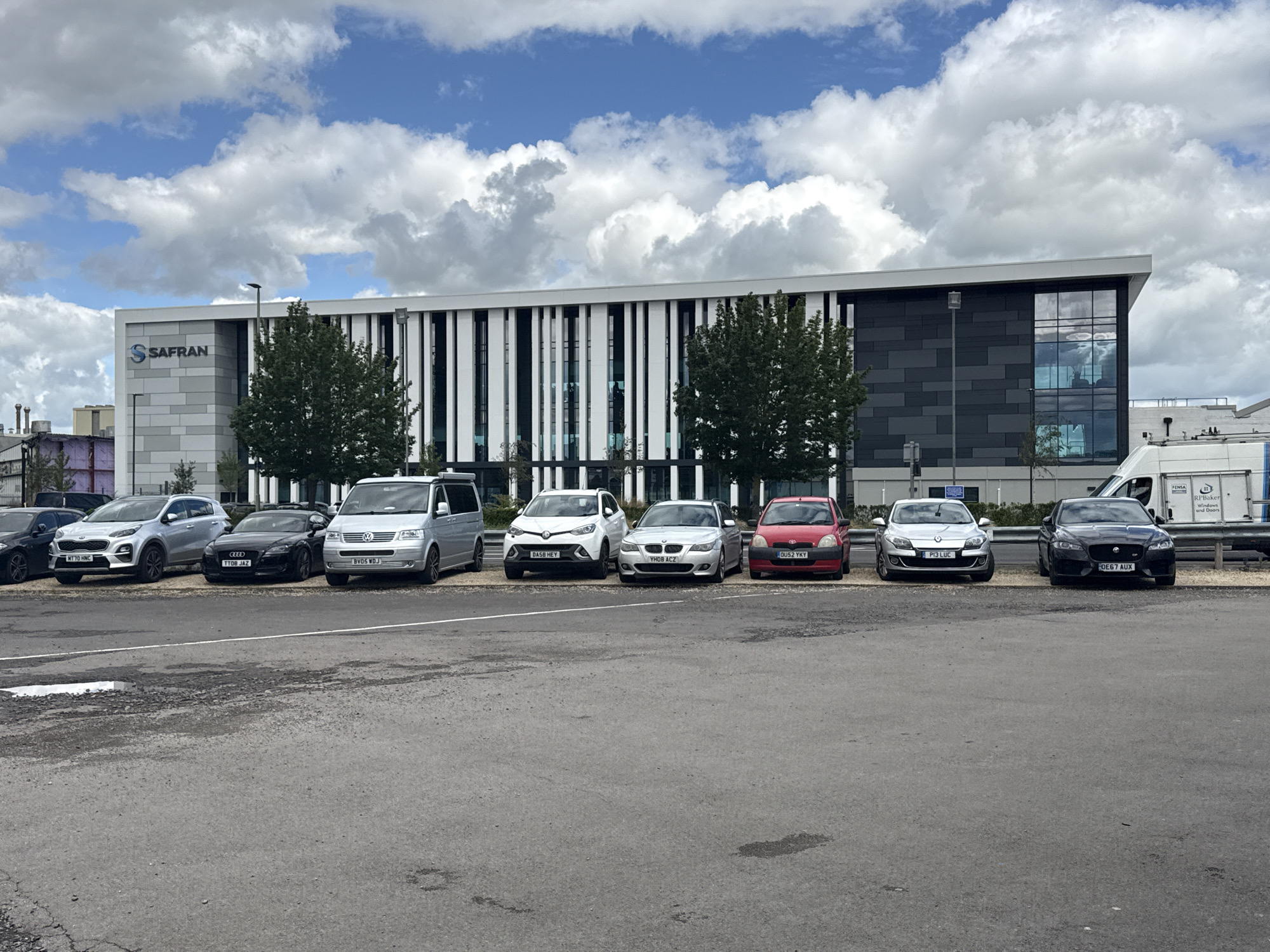 The image shows the Safran building in Tewkesbury, United Kingdom, on a cloudy day. The building is modern with a grey and white facade, featuring vertical white slats and dark grey cladding. A prominent "SAFRAN" logo is visible on the left side of the building. In front of the building, a row of parked cars and a van are visible. These include a silver Kia Sportage, a black Audi TT, a silver Volkswagen Transporter van, a white Suzuki Vitara, a silver BMW 5 Series, a red Ford Fiesta, a silver Renault Megane, and a black Jaguar XF. The ground in the foreground is asphalt with some cracks and puddles. Trees are planted along the front of the building. A white van with "RP Baker" signage is parked on the right side of the image.
